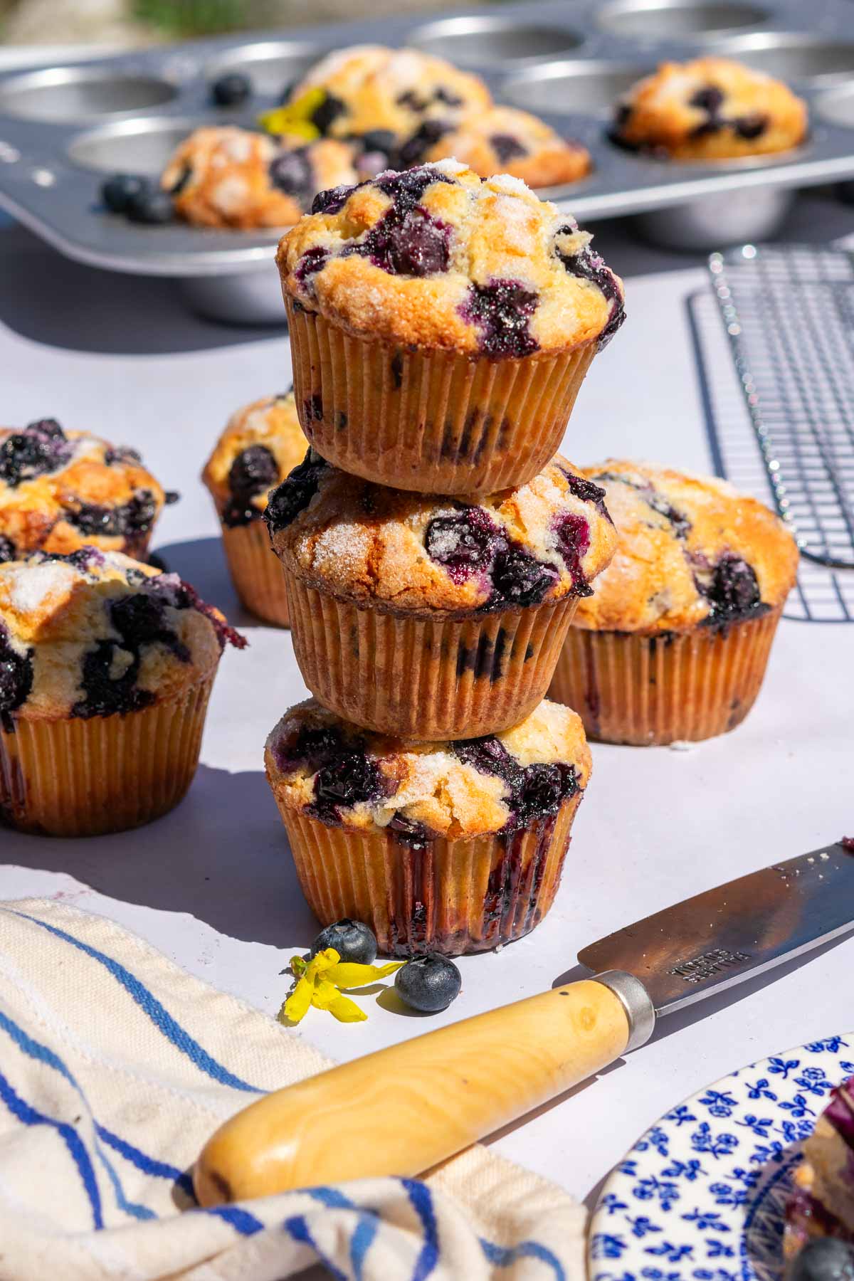 Three sourdough blueberry yogurt muffins stacked on top of each other with a knife, fresh blueberries, a towel, and cooling rack.