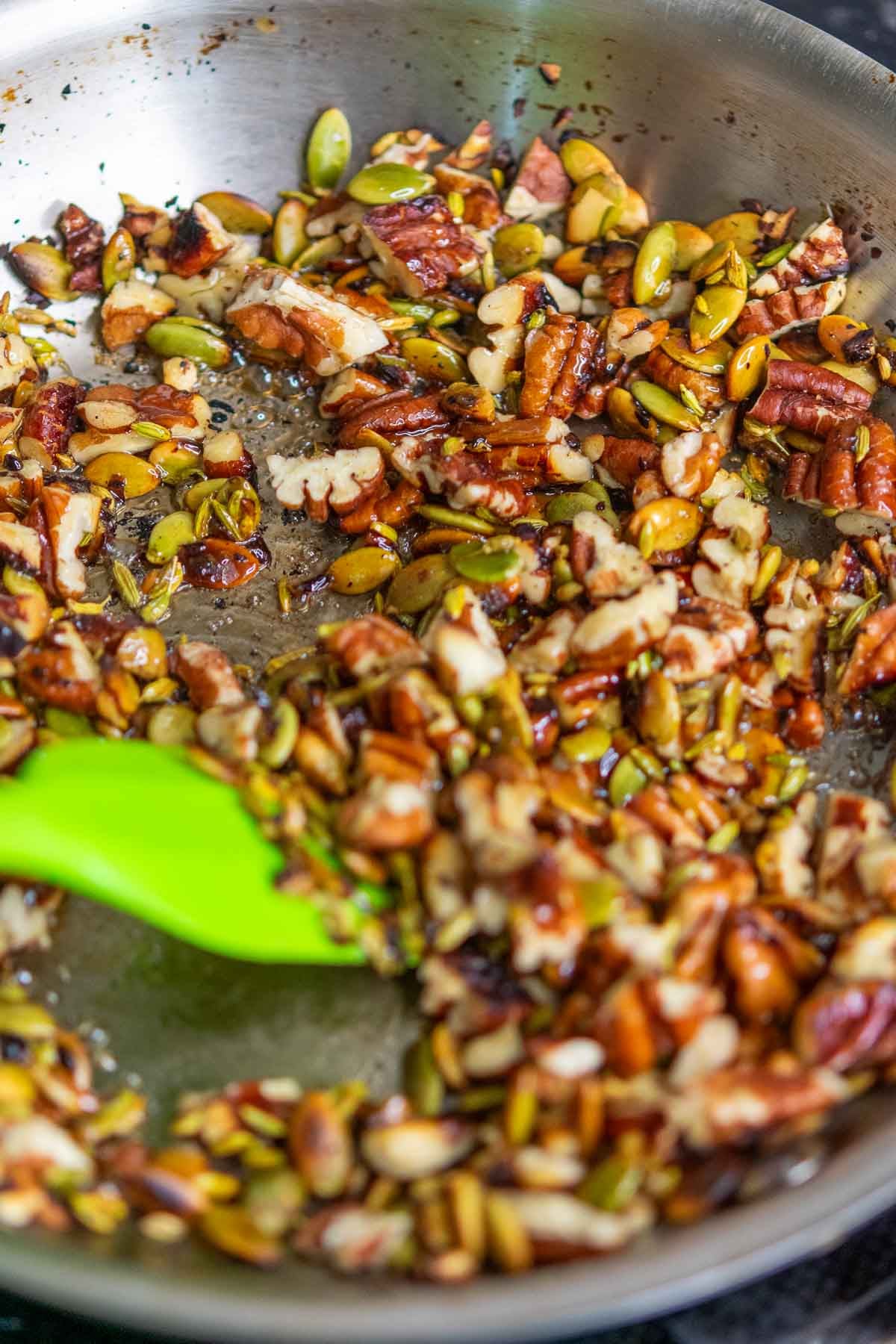 Stirring Maple syrup into skillet with toasted pecans, pumpkin seeds, and fennel seeds in a skillet.