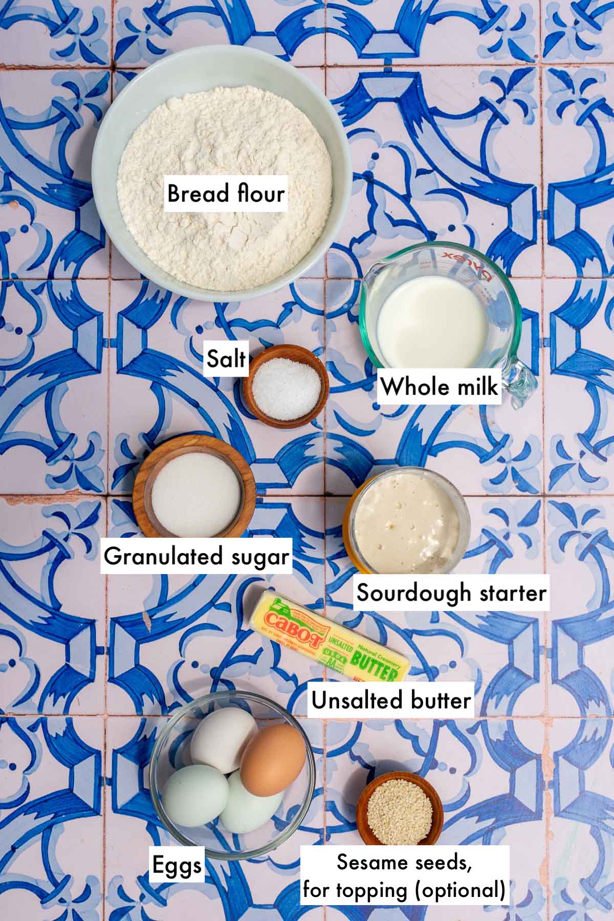 Labeled bowls of ingredients needed for sourdough brioche hamburger buns including bread flour, salt, sugar, sourdough starter, butter, eggs, sesame seeds, and whole milk.