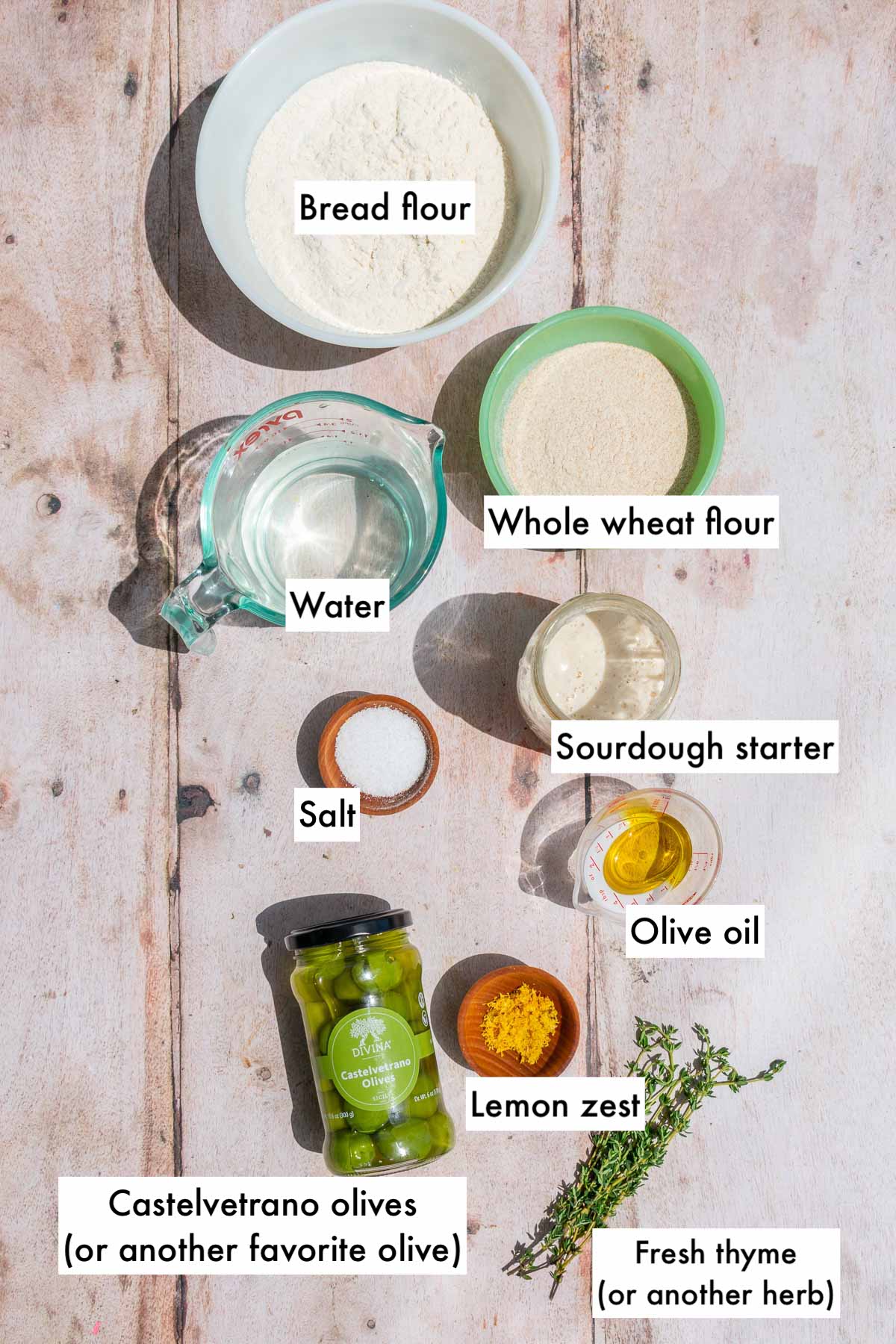 Labeled bowls of ingredients needed for olive sourdough bread including bread flour, whole wheat flour, Castelvetrano olives, lemon zest, thyme, water, sourdough starter, salt, and olive oil.