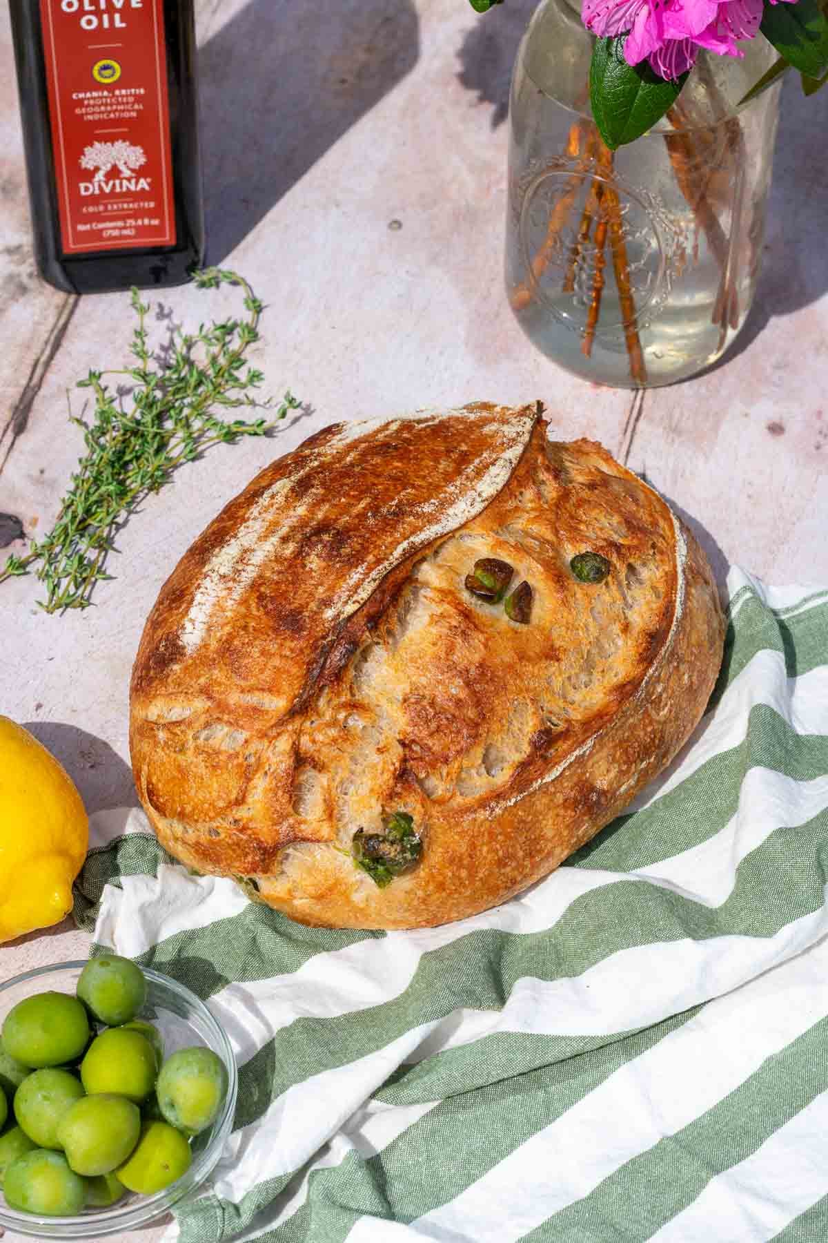 Loaf of olive sourdough bread with a bowl of castelvetrano olives, a lemon, thyme leaves, and olive oil.