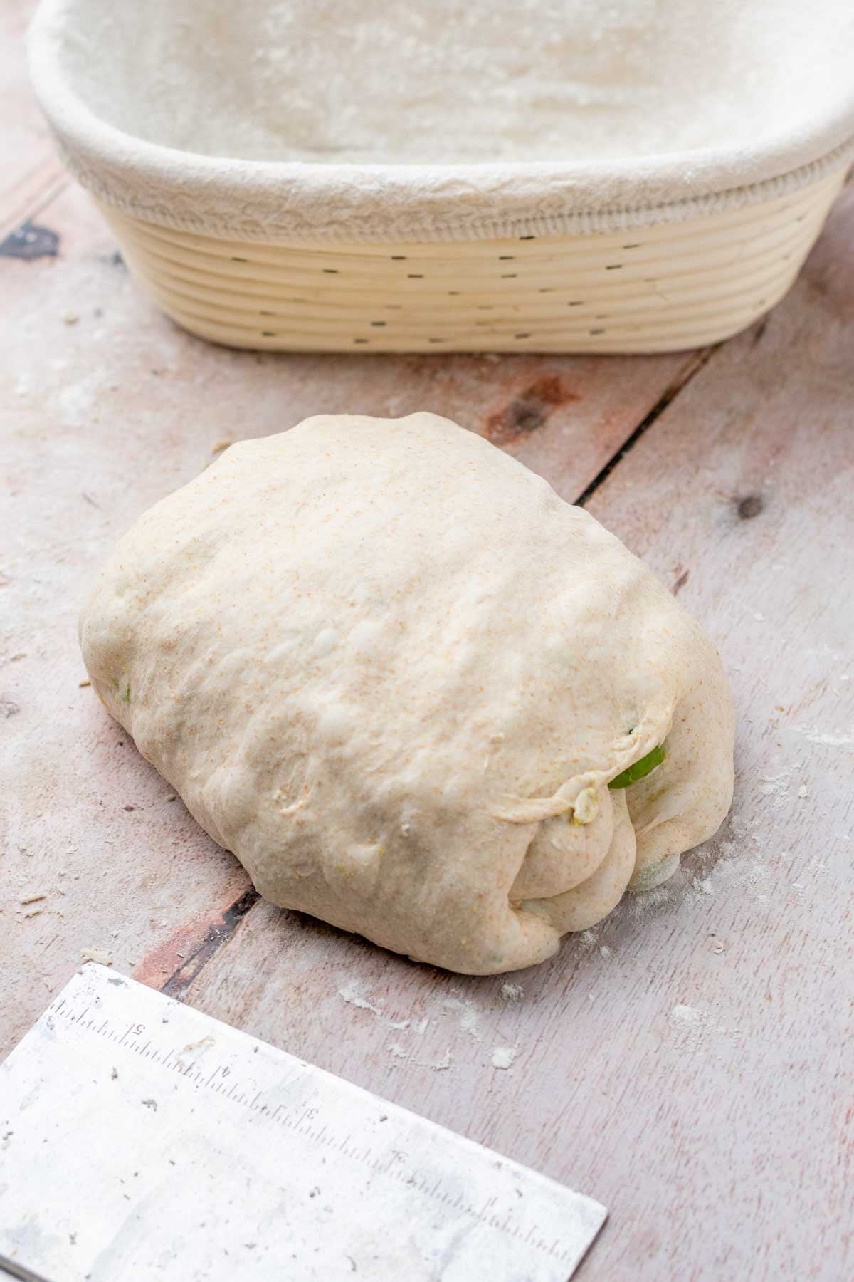 Shaped olive sourdough bread on a counter with a banneton and a bench scraper.