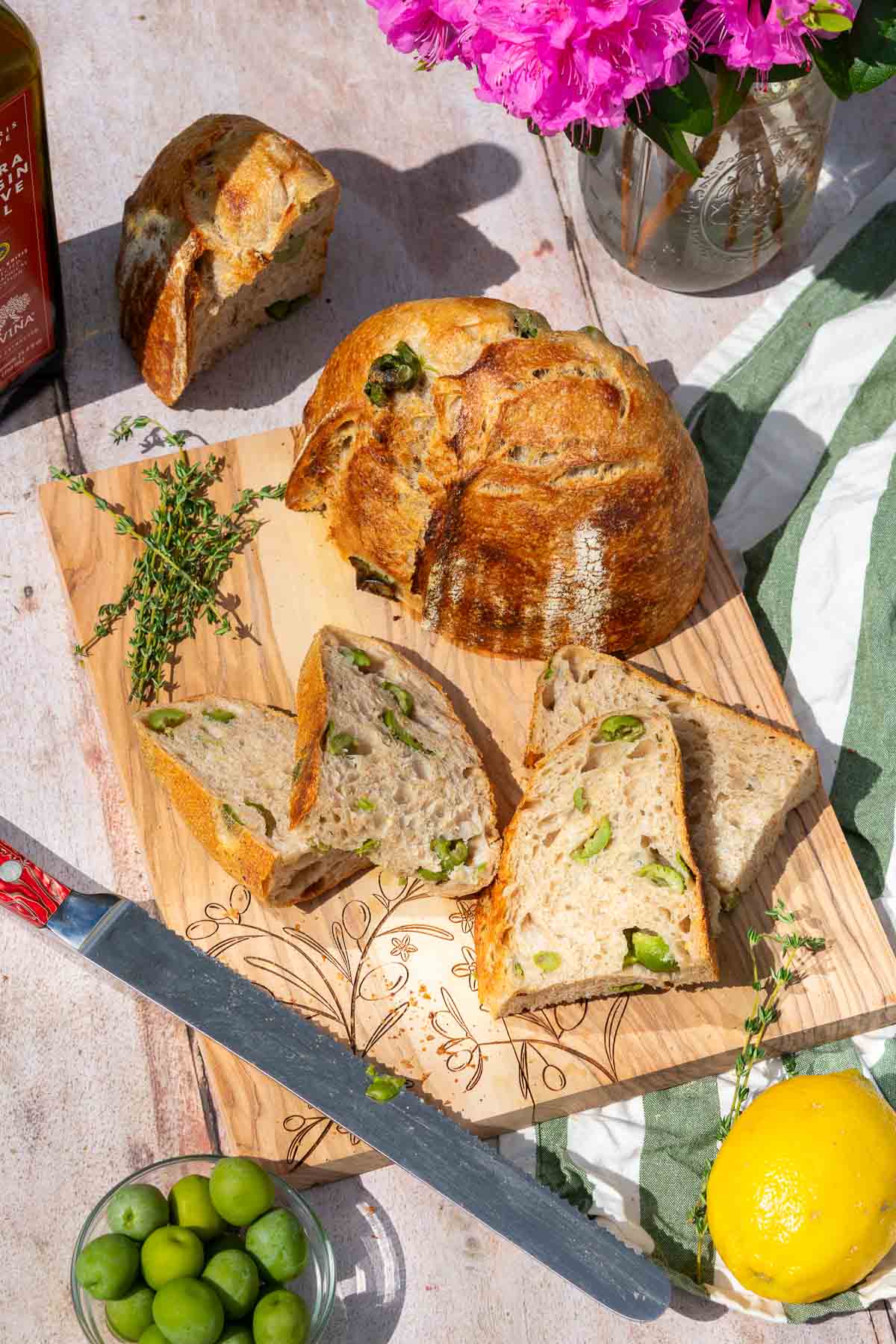 Sliced olive sourdough bread on a cutting board with a bread knife, a bowl of olives, lemon, and thyme.