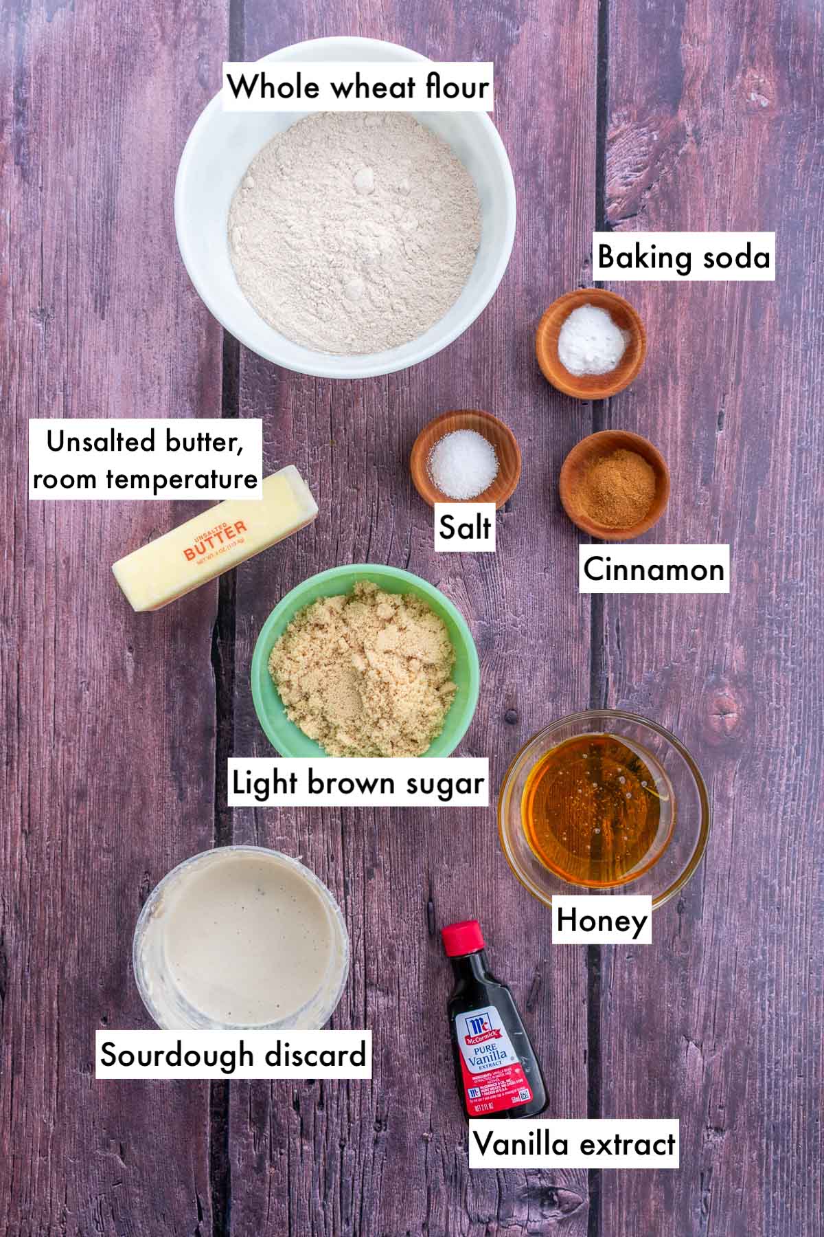 Bowls of labeled ingredients needed for sourdough graham crackers including whole wheat flour, salt, baking soda, butter, cinnamon, light brown sugar, honey, sourdough discard, and vanilla extract.