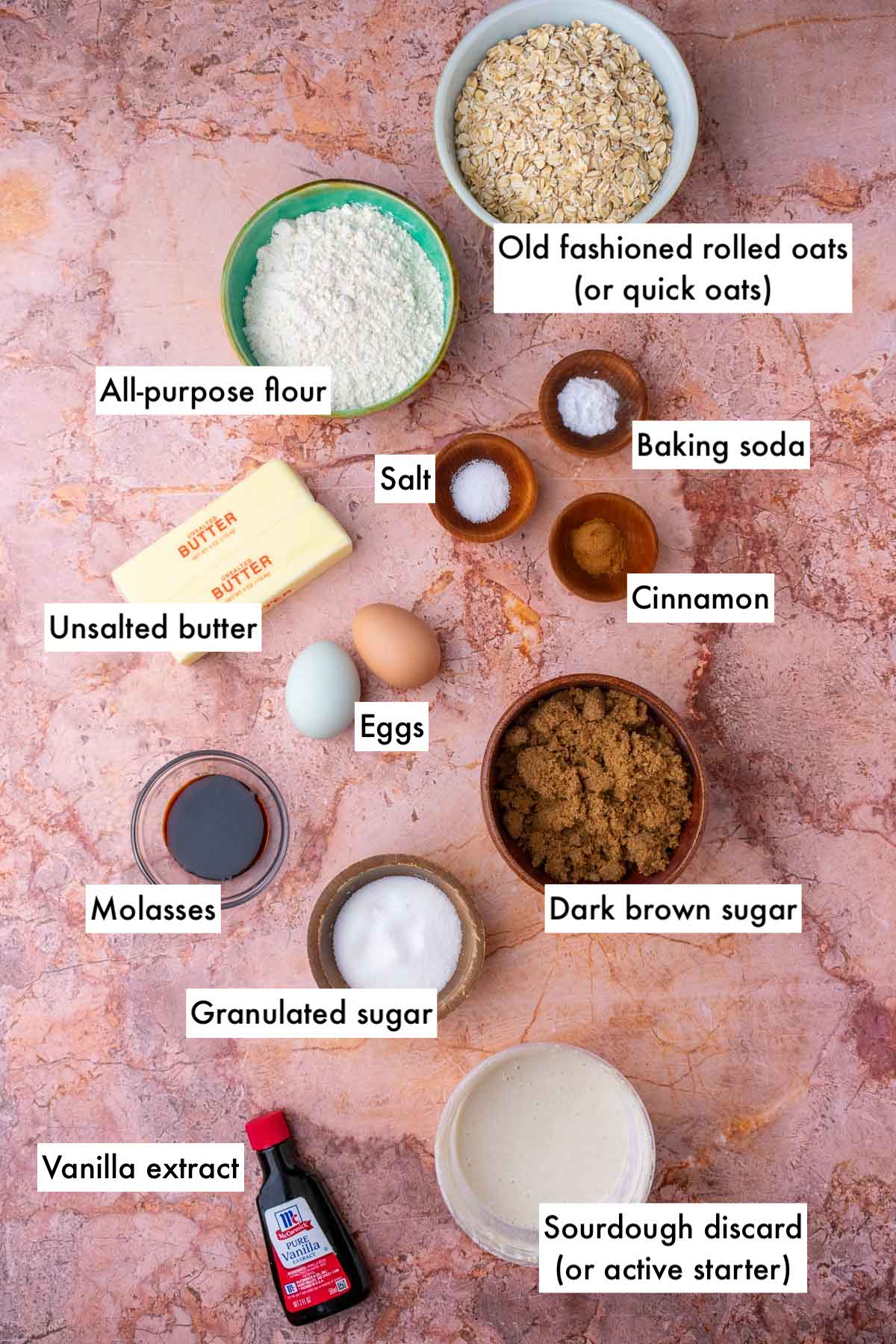 Labeled ingredients needed for sourdough oatmeal cream pies including oats, flour, cinnamon, baking soda, salt, unsalted butter, eggs, brown sugar, granulated sugar, molasses, sourdough discard, and vanilla.
