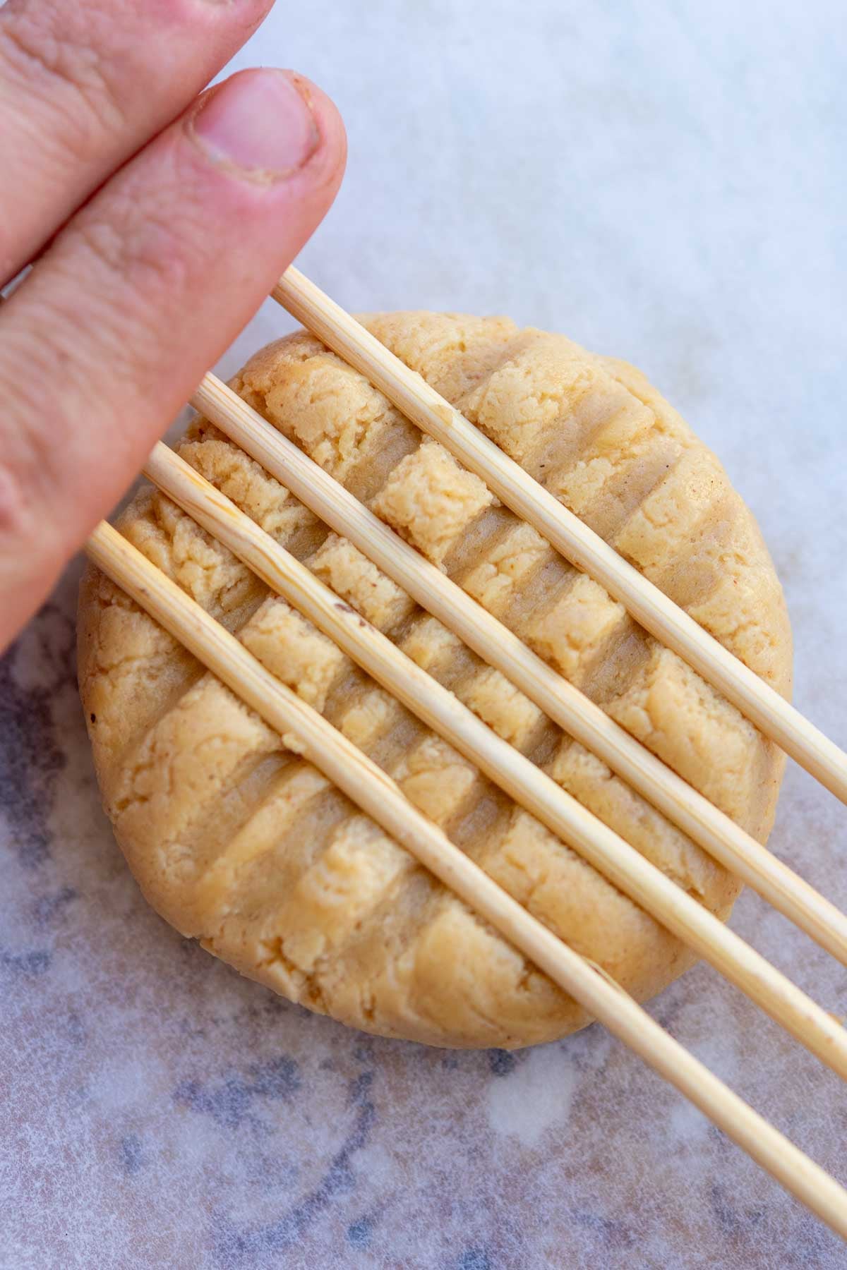 Hand pressing down on sourdough peanut butter cookie with wooden dowels to make crisscross pattern.