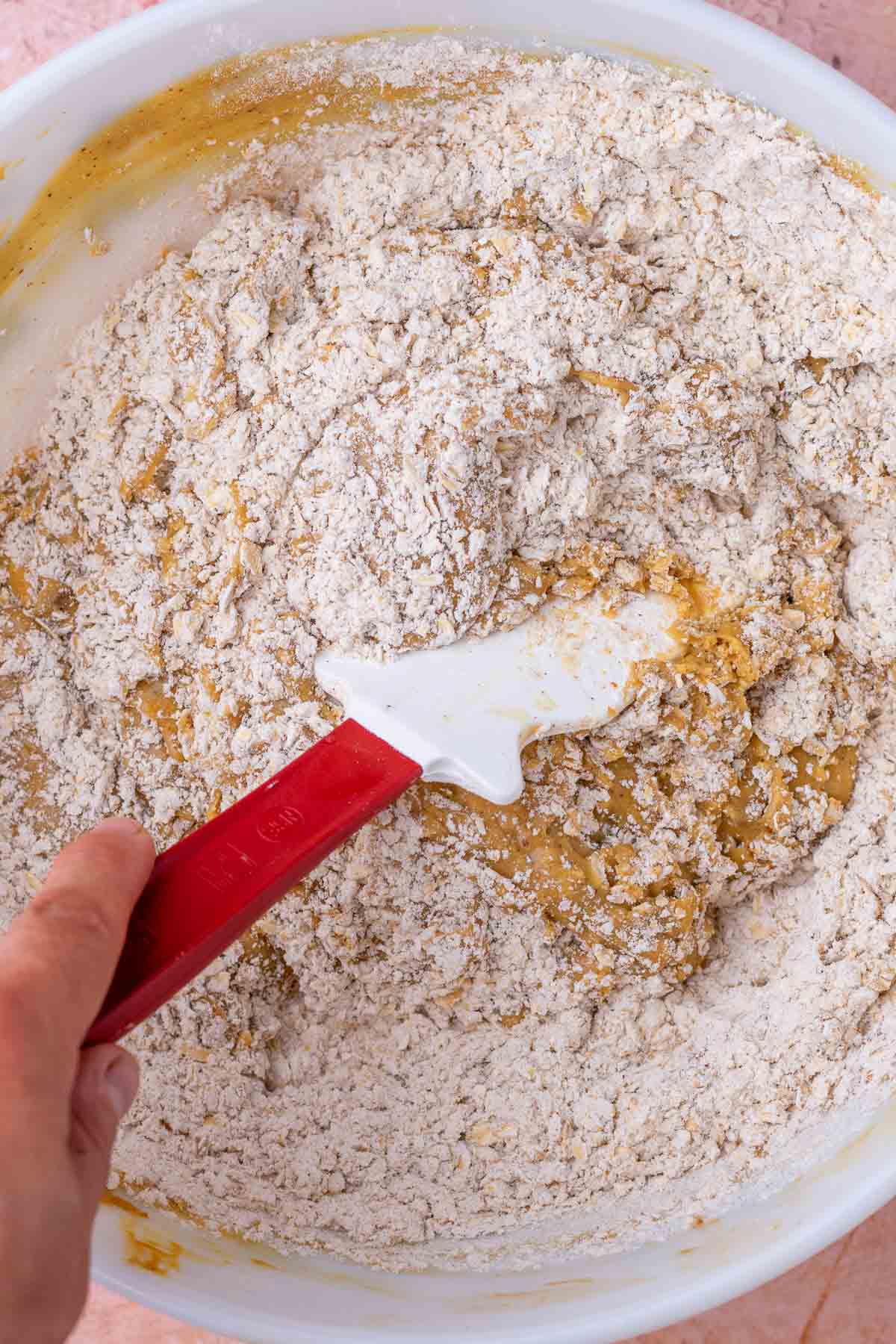 Spatula mixing dry ingredients into bowl of wet ingredients for sourdough oatmeal cookies.