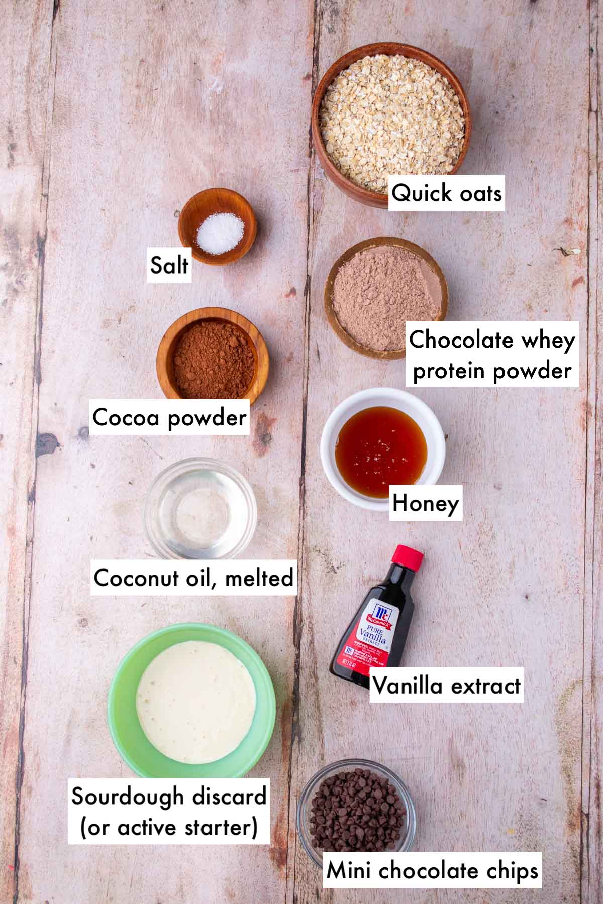 Bowls of ingredients needed for sourdough chocolate protein granola bar including quick oats, salt, cocoa powder, whey protein powder, vanilla extract, sourdough discard, honey, coconut oil, and mini chocolate chips.