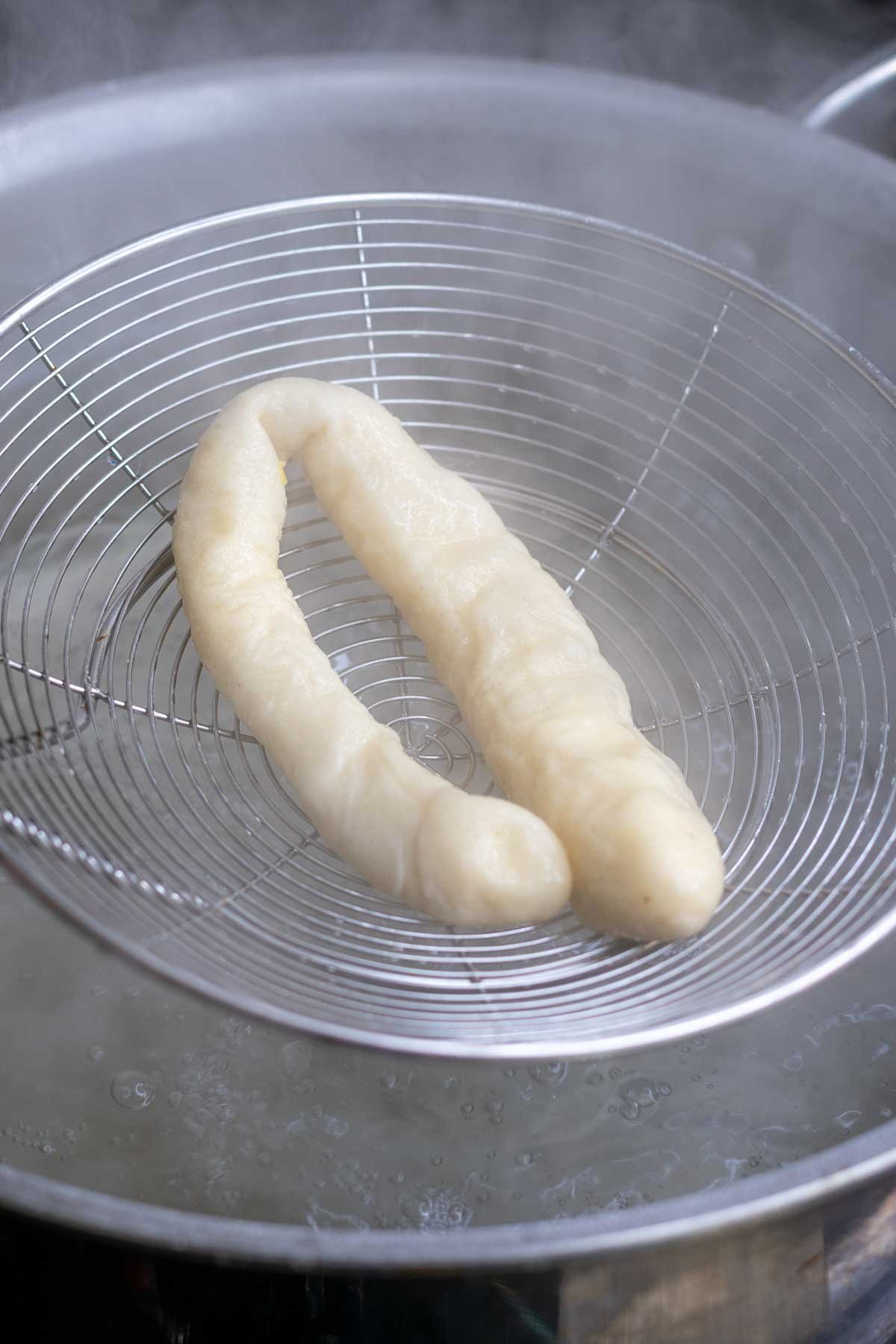 Lifting a boiled sourdough pretzel rod with a spider strainer from a pot.