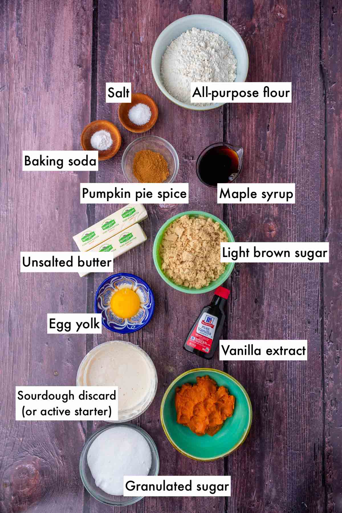 Labeled bowls of ingredients needed for sourdough pumpkin cookies including all-purpose flour, baking soda, butter, brown sugar, pumpkin puree, sourdough discard, egg yolk, vanilla extract, granulated sugar, maple syrup, spices, and salt.