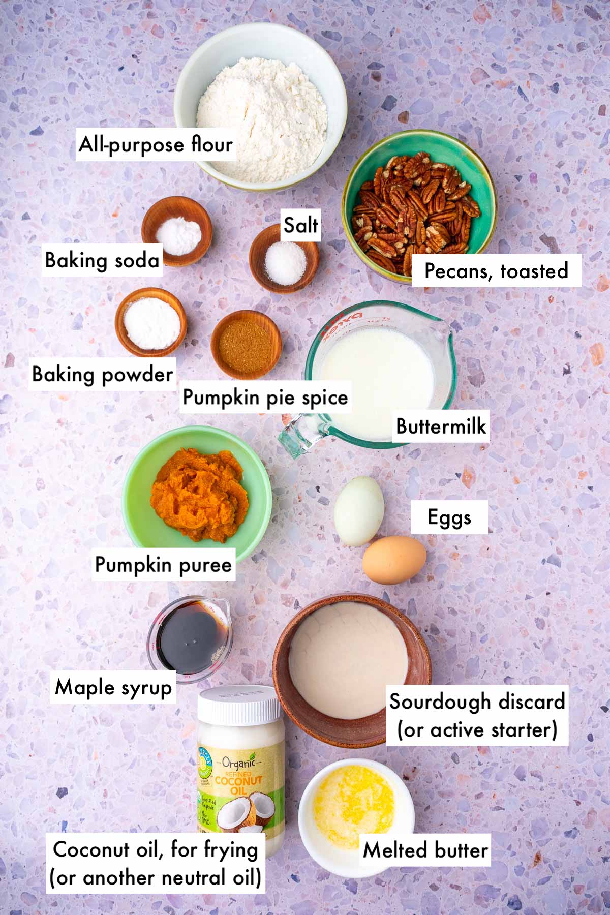 Labeled bowls of ingredients needed for sourdough pumpkin pancakes including all-purpose flour, salt, baking powder and soda, pumpkin puree, sourdough discard, eggs, maple syrup, oil for frying, butter, and pumpkin pie spice.
