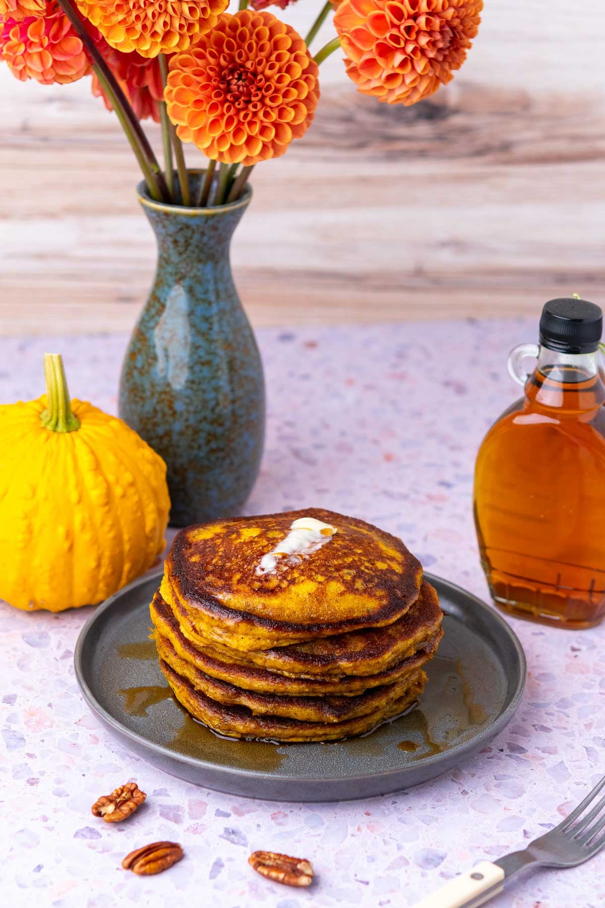A stack of sourdough pumpkin pancakes with butter and maple syrup and flowers and a pumpkin in the background.