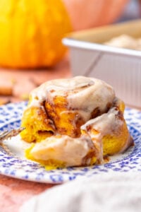 Bite taken out of a sourdough pumpkin cinnamon roll with chai cream cheese frosting on a plate and a pumpkin in the background.