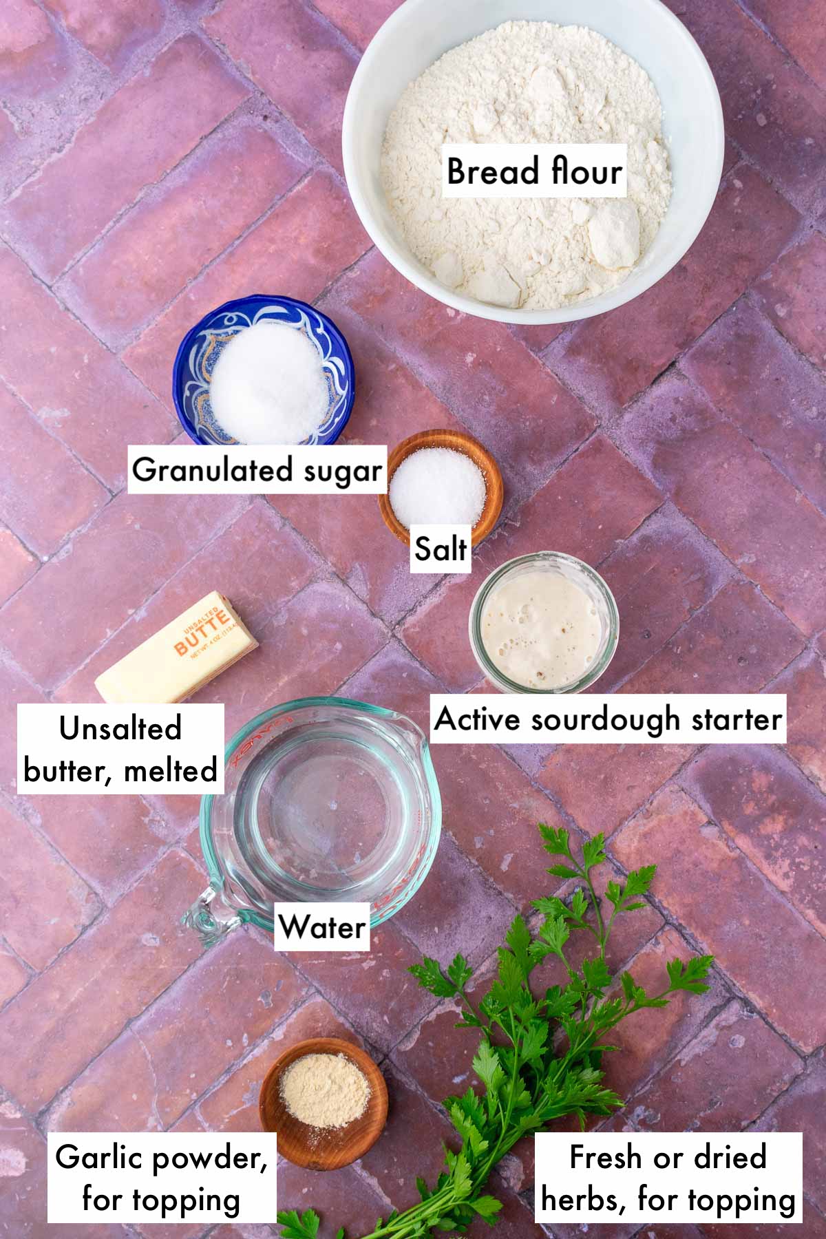 Labeled bowls of ingredients needed for sourdough breadsticks, including bread flour, salt, sugar, butter, water, active sourdough starter, garlic powder, and parsley.