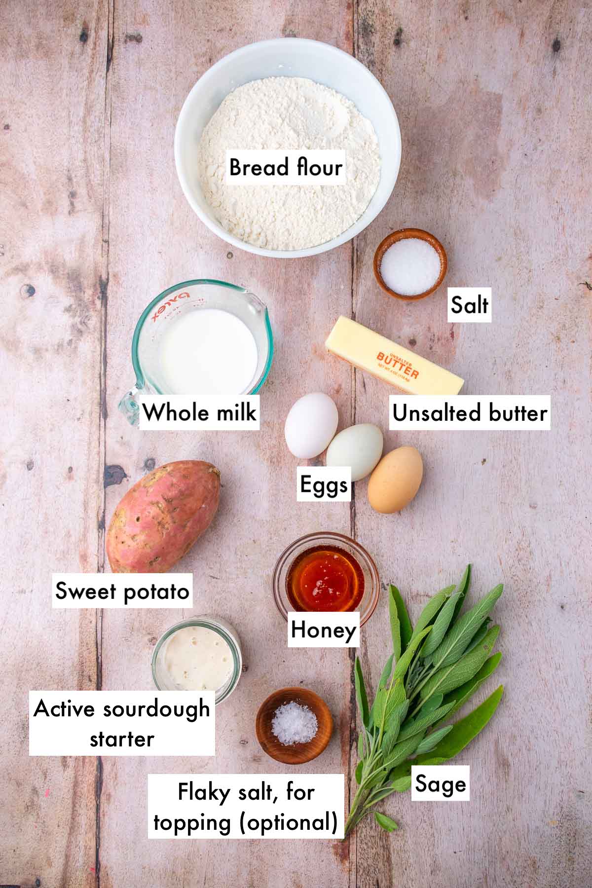 Labeled bowls of ingredients needed for sourdough sweet potato dinner rolls, including bread flour, salt, butter, whole milk, eggs, sage, sweet potato, sourdough starter, honey, and flaky salt.