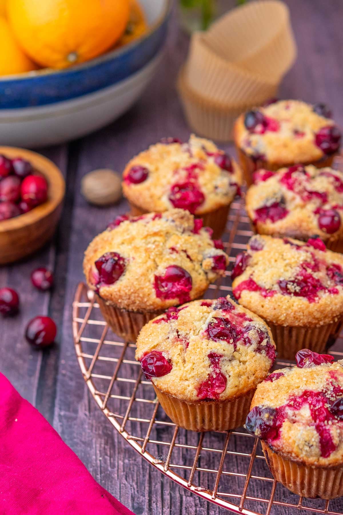 Sourdough cranberry muffins on a wire rack with muffin liners, fresh cranberries, and bowl of oranges in the background.