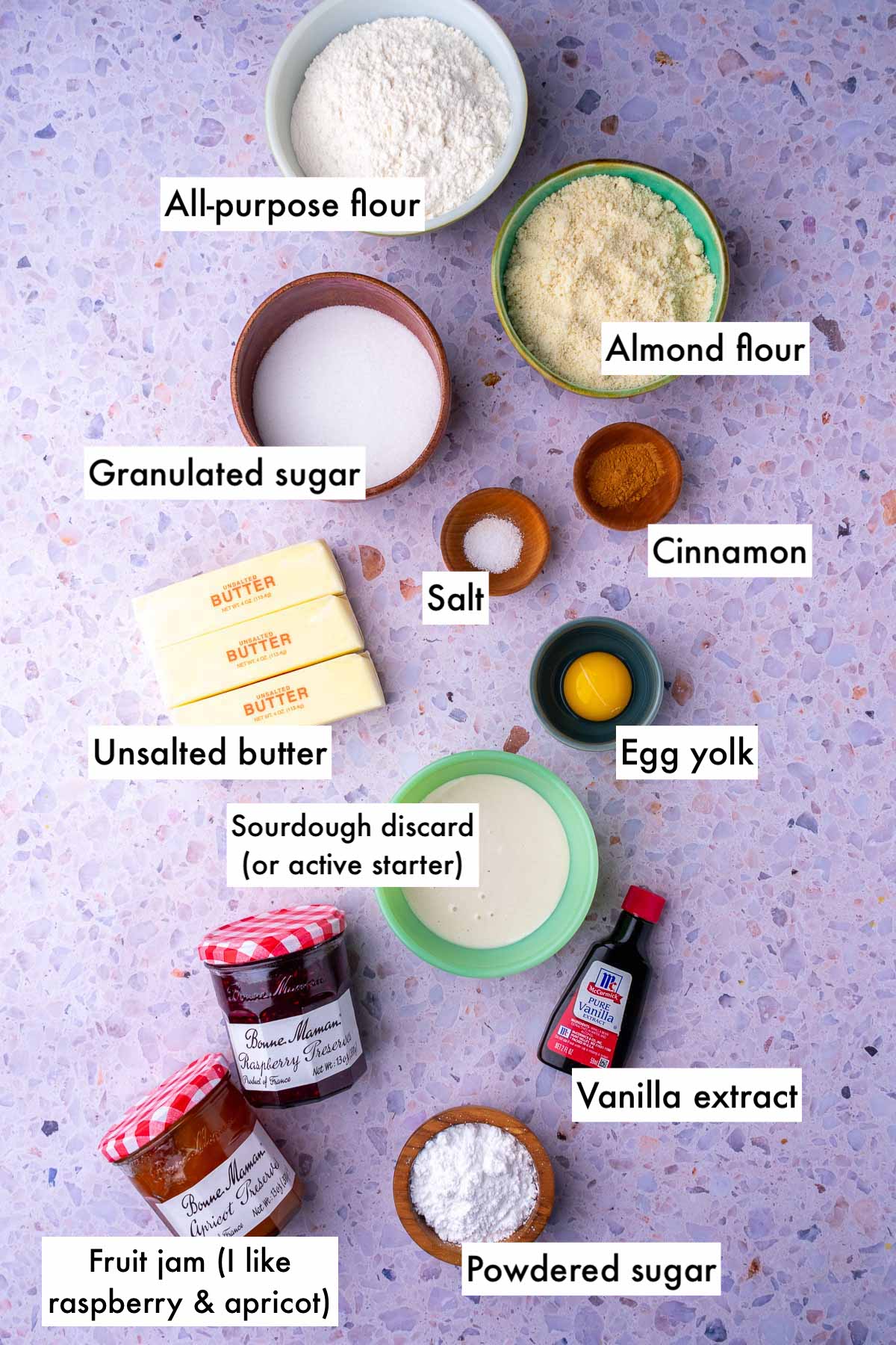Labeled bowls of ingredients needed for sourdough linzer cookies including all-purpose flour, almond flour, sugar, butter, cinnamon, salt, egg yolk, sourdough discard, vanilla extract, powdered sugar, and fruit jams.