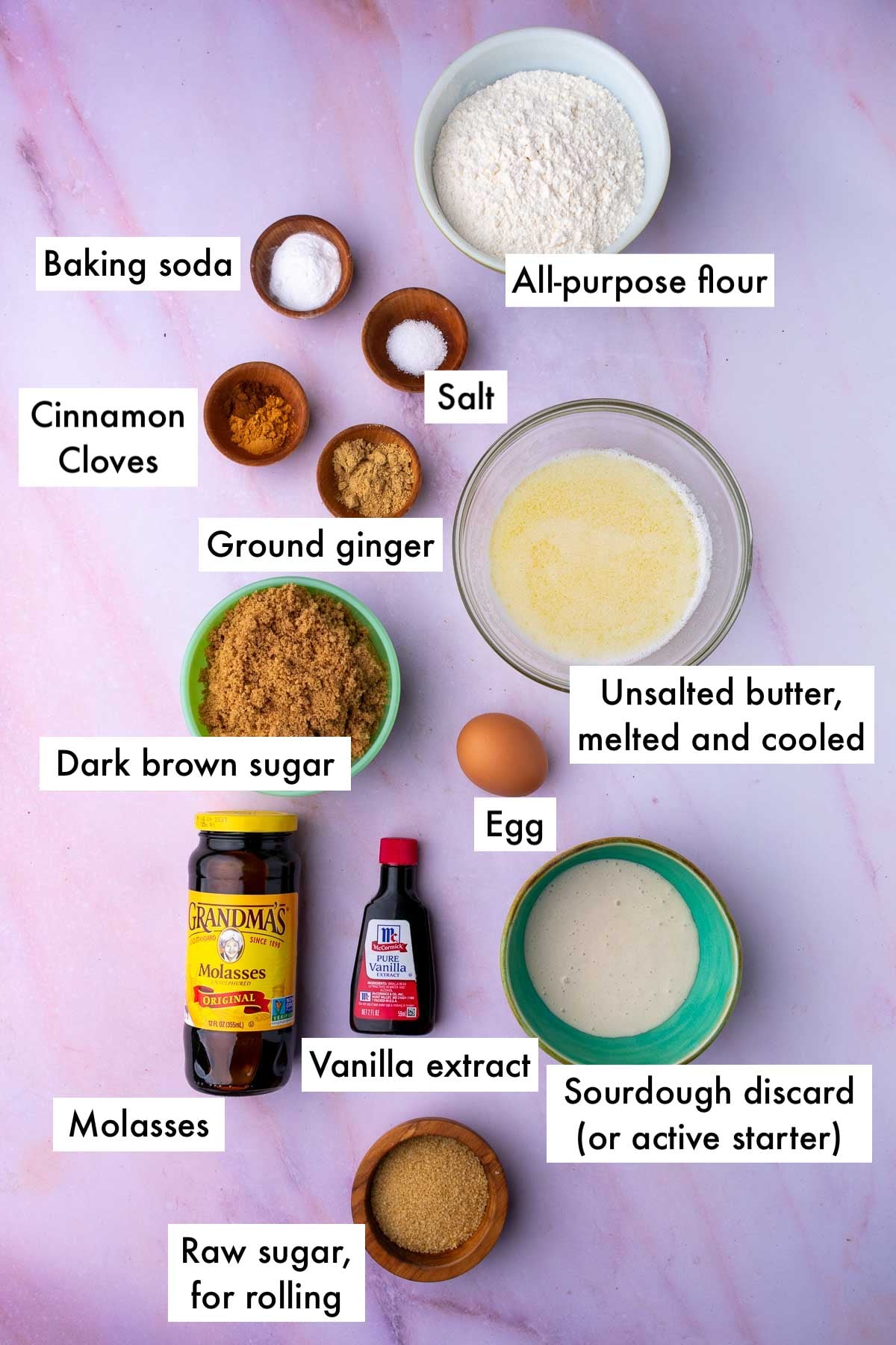Labeled bowls of ingredients for Sourdough ginger molasses cookies, including all-purpose flour, salt, baking soda, melted butter, cinnamon, cloves, ginger, dark brown sugar, egg, sourdough discard, vanilla extract, molasses, and raw sugar.