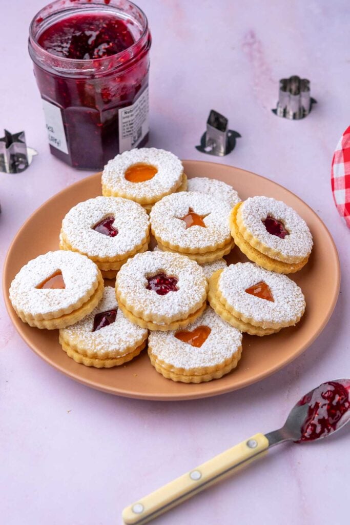 Plate of sourdough linzer cookies with a jar of raspberry jam and cookie cut outs.