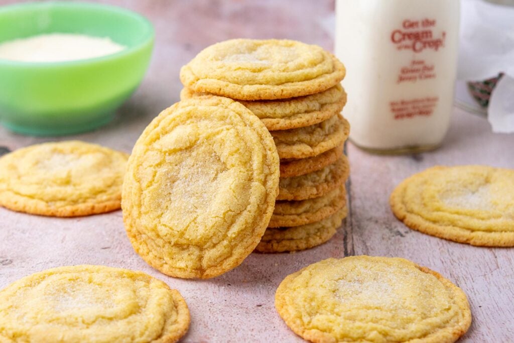 A stack of sourdough sugar cookies with a bowl of sugar and a glass of milk in the background.