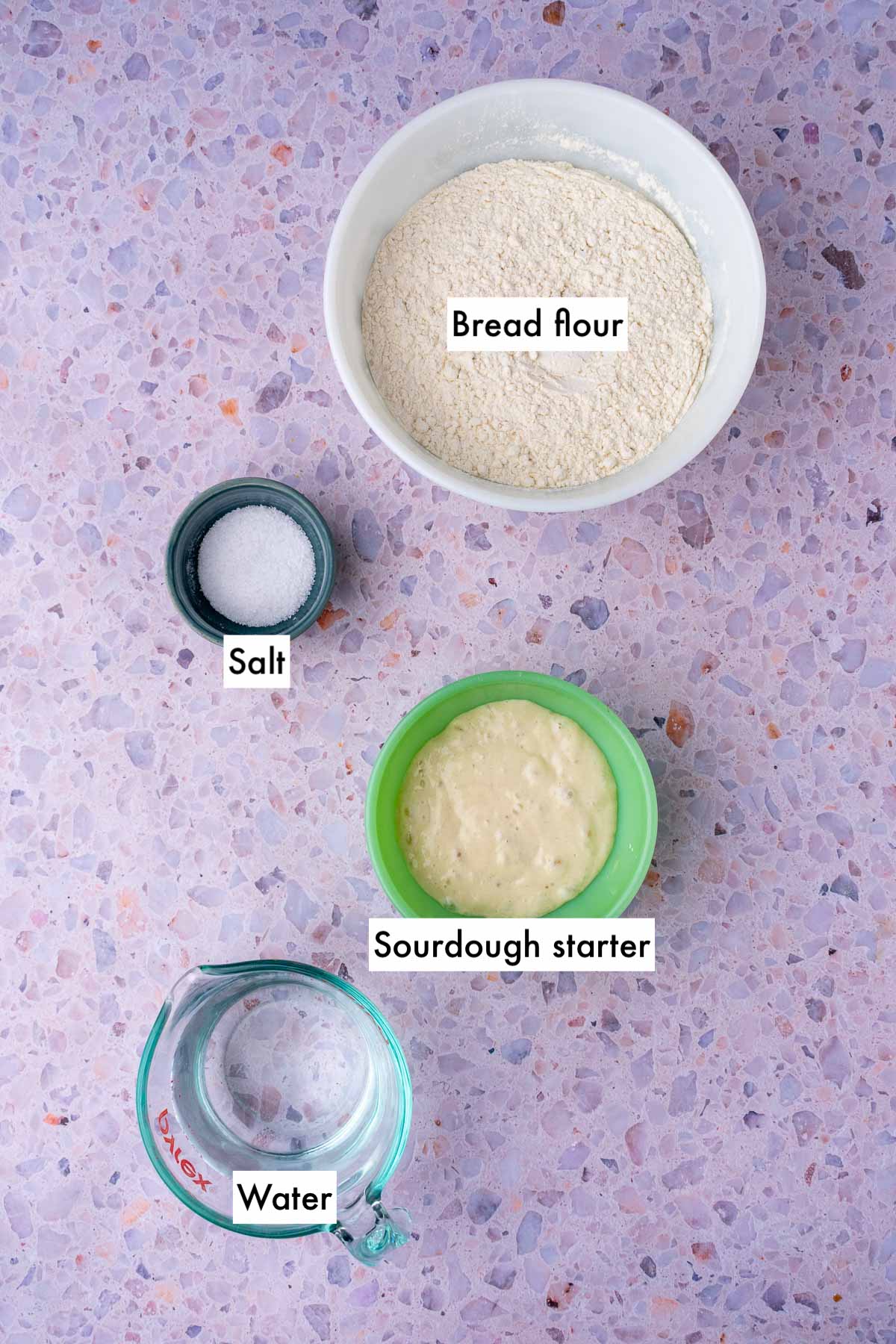 Labeled bowls of ingredients needed for sourdough baguettes including bread flour, salt, sourdough starter, and water.