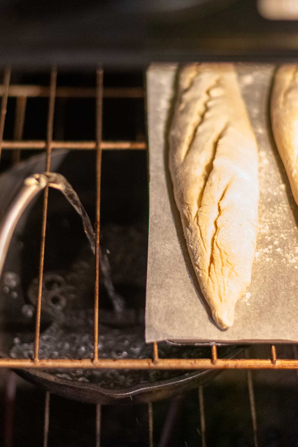 Pouring boiling water into a cast-iron skillet to create steam in oven with sourdough baguettes on a baking steel.