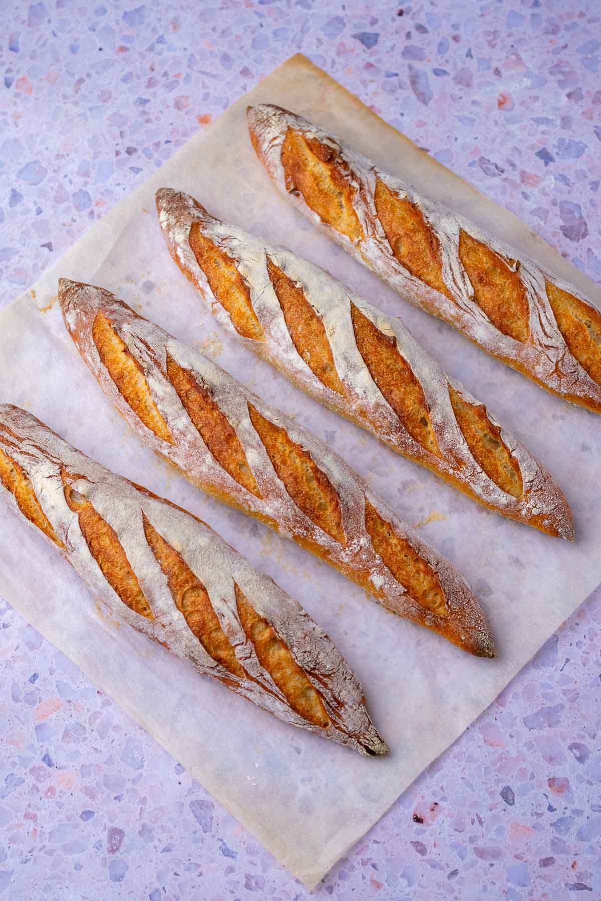 Four baked sourdough baguettes on a baking steel with parchment paper.