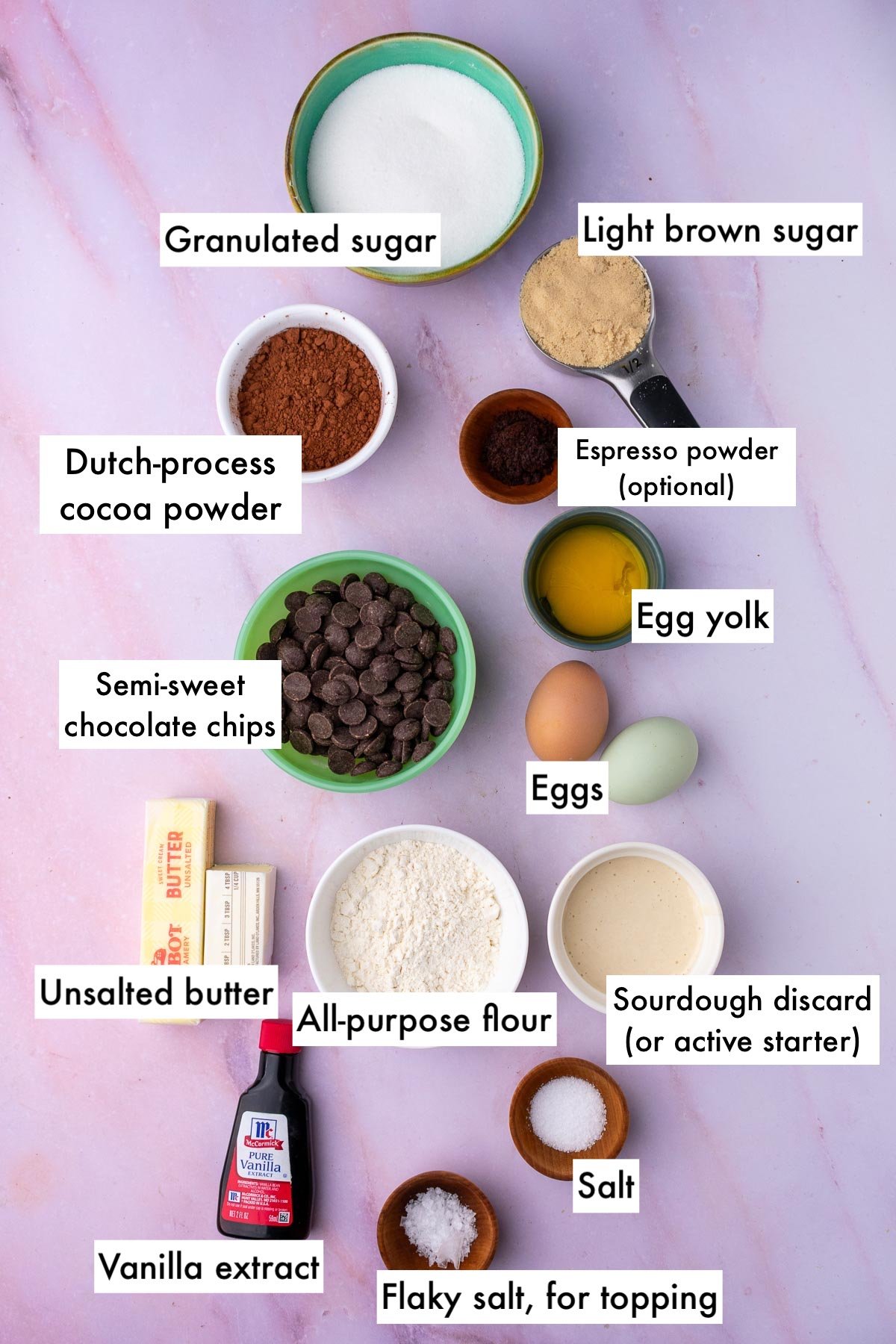 Labeled bowls of ingredients needed for sourdough brownies including sugar, light brown sugar, espresso powder, egg yolk, eggs, sourdough discard, chocolate chips, cocoa powder, vanilla extract, unsalted butter, salt, flaky salt, and all-purpose flour.