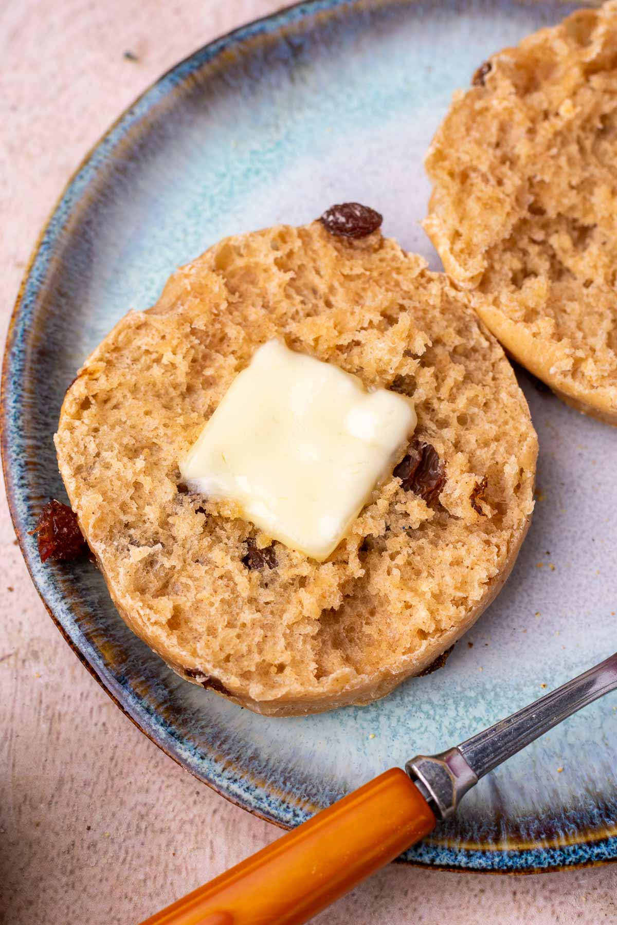Close up of a sourdough cinnamon raisin english muffin cut in half with butter on a plate.