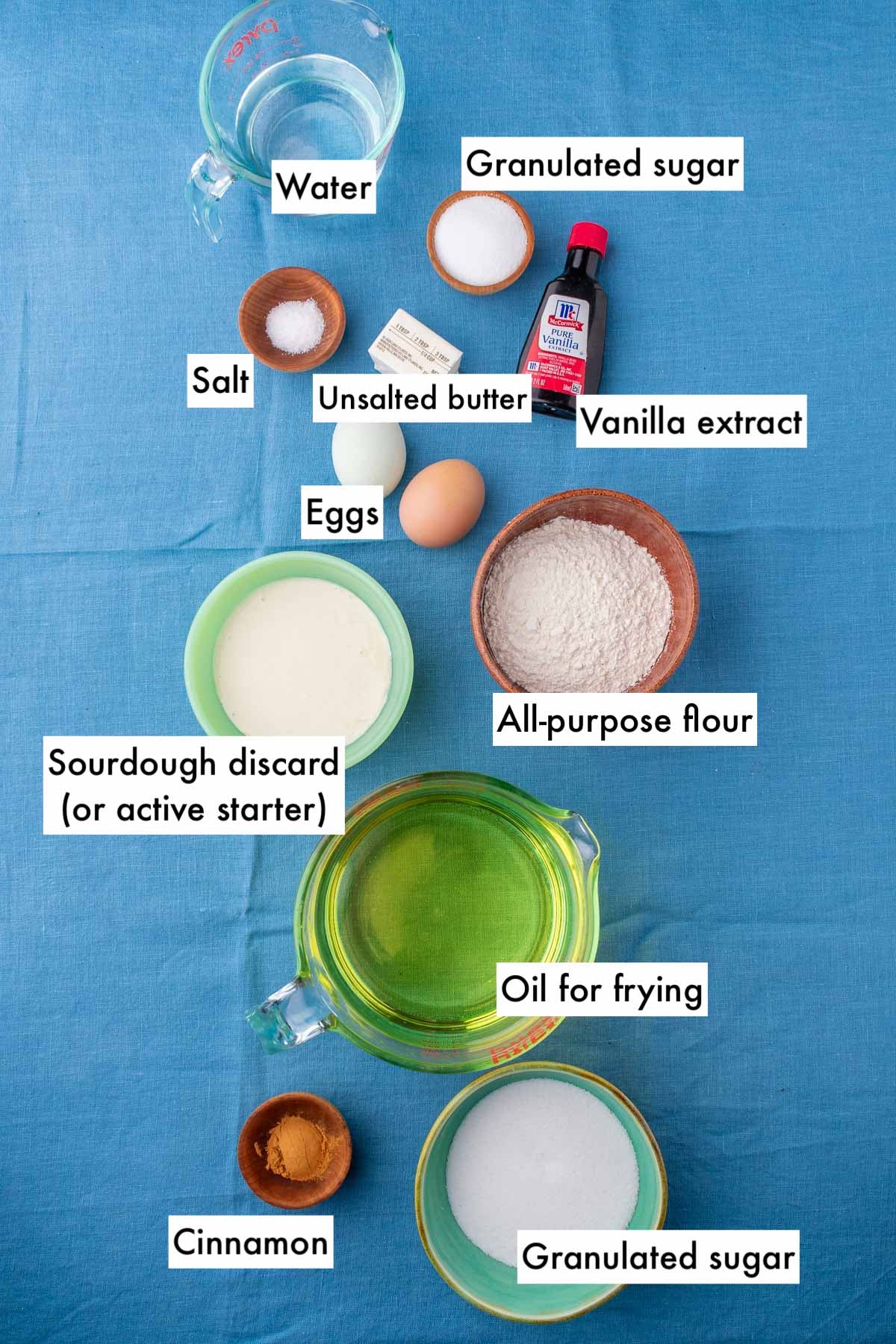 Labeled bowls of ingredients needed for sourdough churros including all-purpose flour, water, sourdough discard, granulated sugar, cinnamon, vanilla, oil, eggs, salt, and unsalted butter.