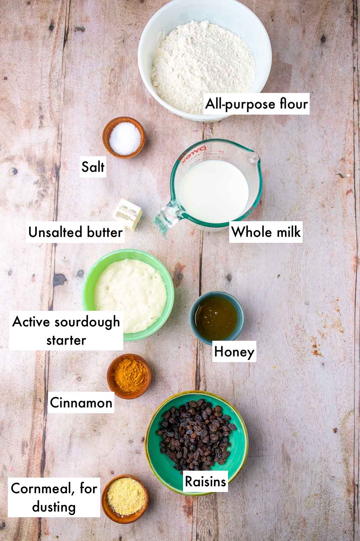 Labeled bowls of ingredients needed for sourdough cinnamon raisin english muffins, including all-purpose flour, salt, whole milk, active sourdough starter, raisins, honey, unsalted butter, cinnamon, and cornmeal for dusting.
