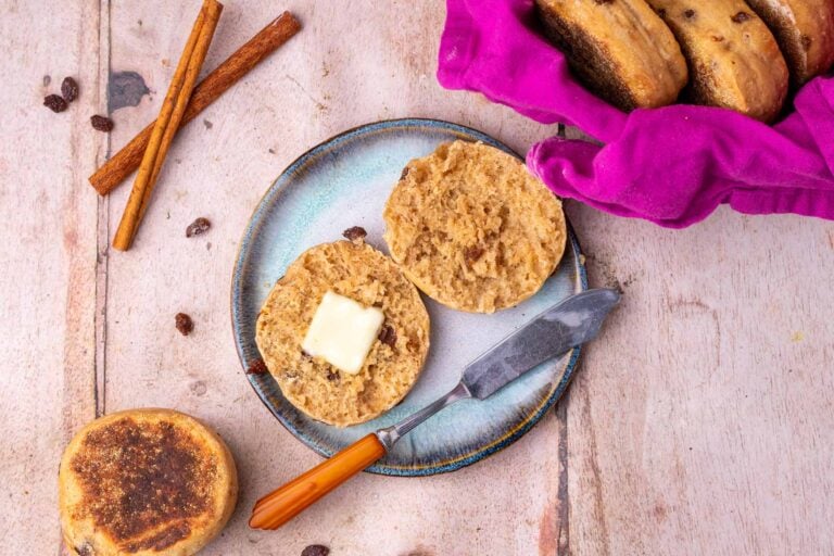 Plate with a sourdough cinanmon raisin english muffin sliced open with butter and a butter knife, cinnamon sticks, raisins, and english muffins to the side.