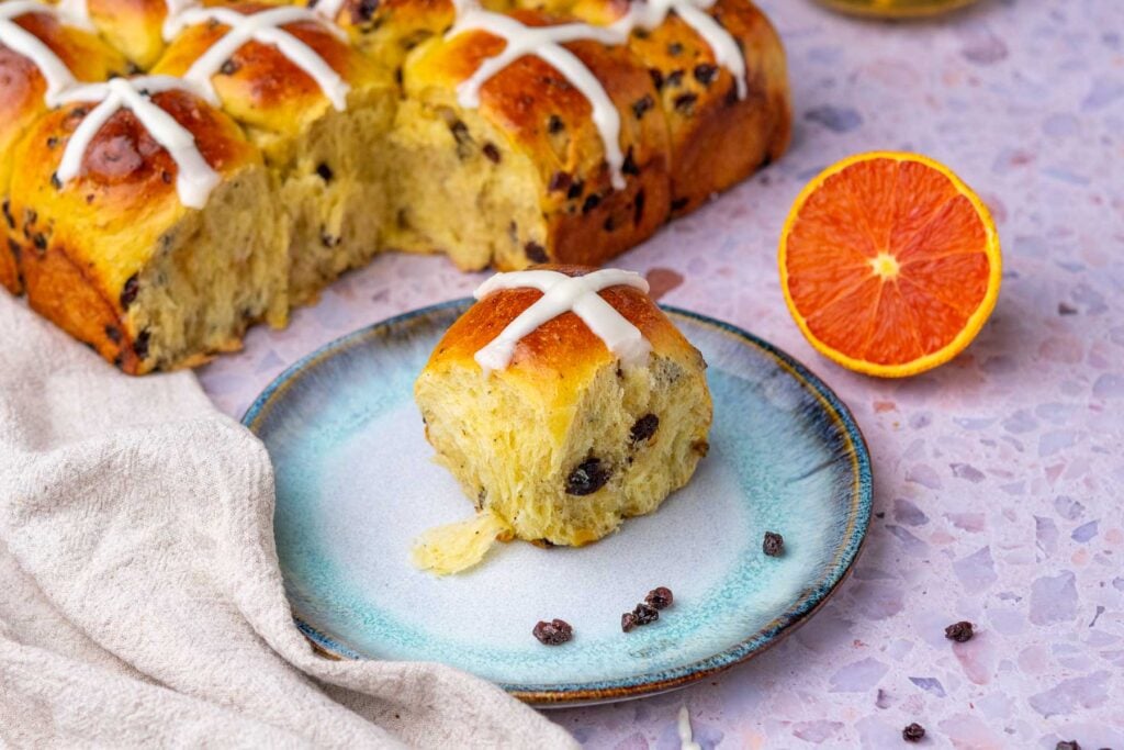 Sourdough hot cross buns on a plate with currants and a sliced orange.