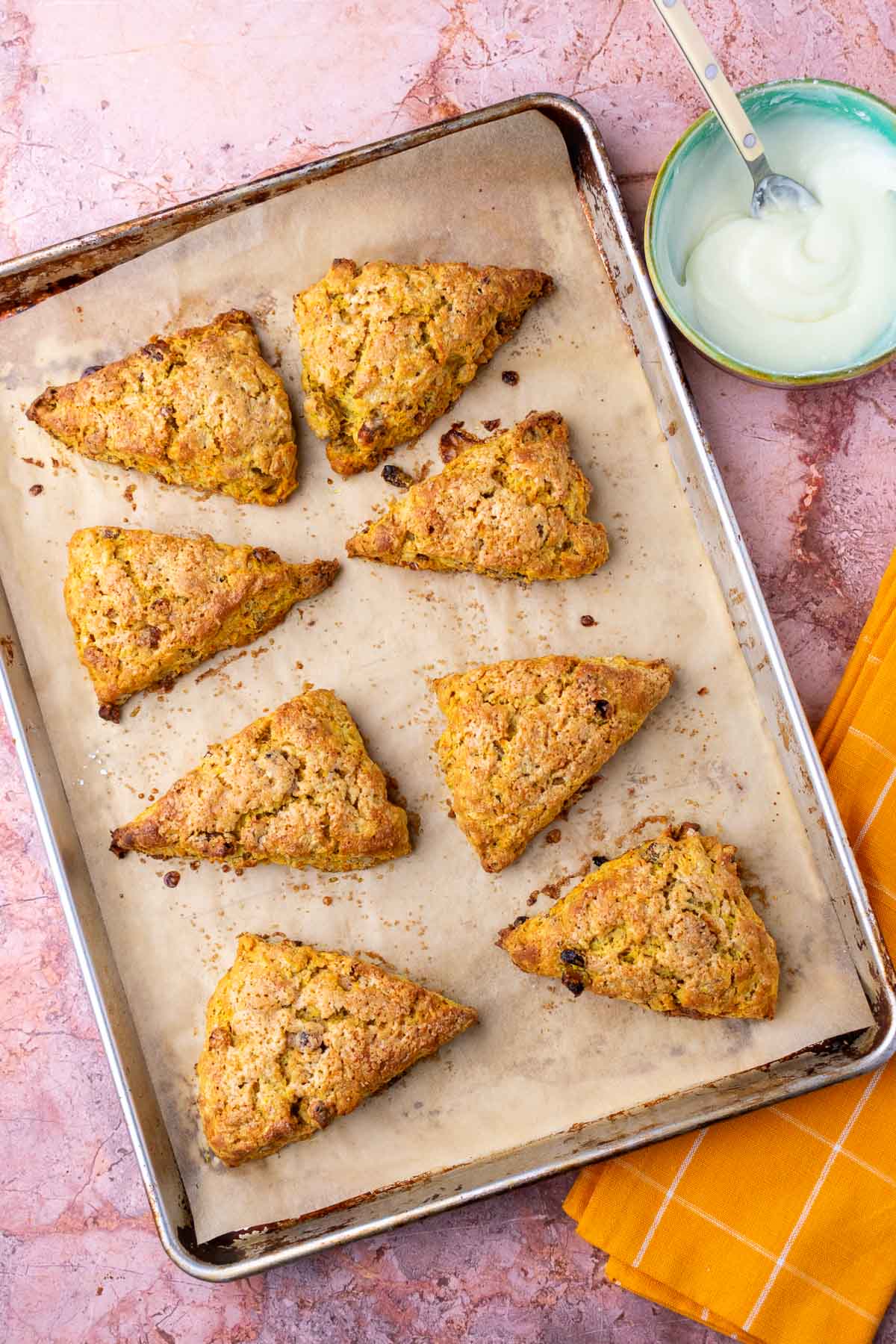 Baked sourdough carrot cake scones on baking sheet with a bowl of cream cheese glaze to the side.