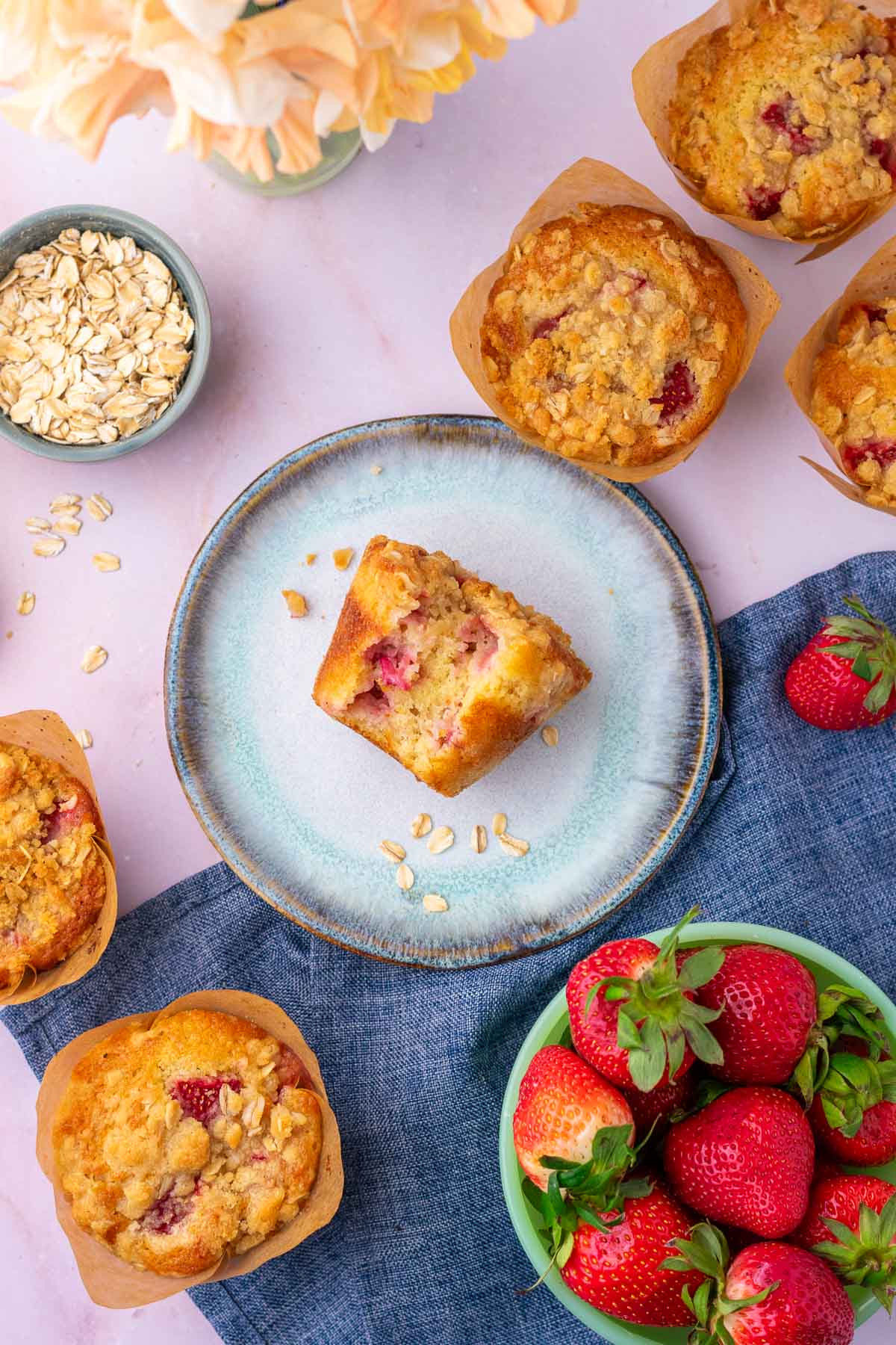 Bite taken out of a Sourdough strawberry muffin on a plate surrounded by more Sourdough strawberry muffins, a bowl of oats, and a bowl of strawberries.