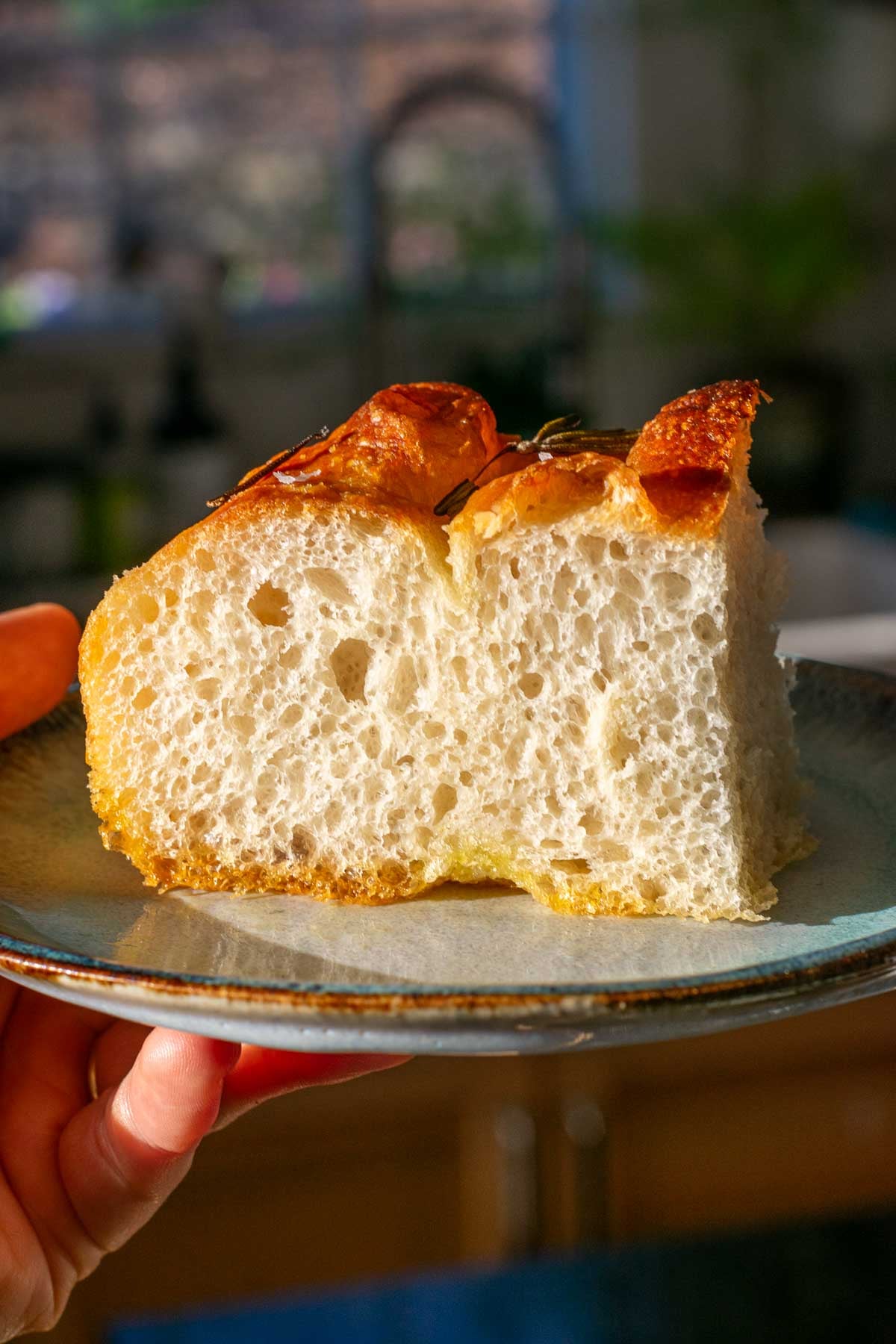 Hand holding a slice of sourdough focaccia with open crumb on a plate.