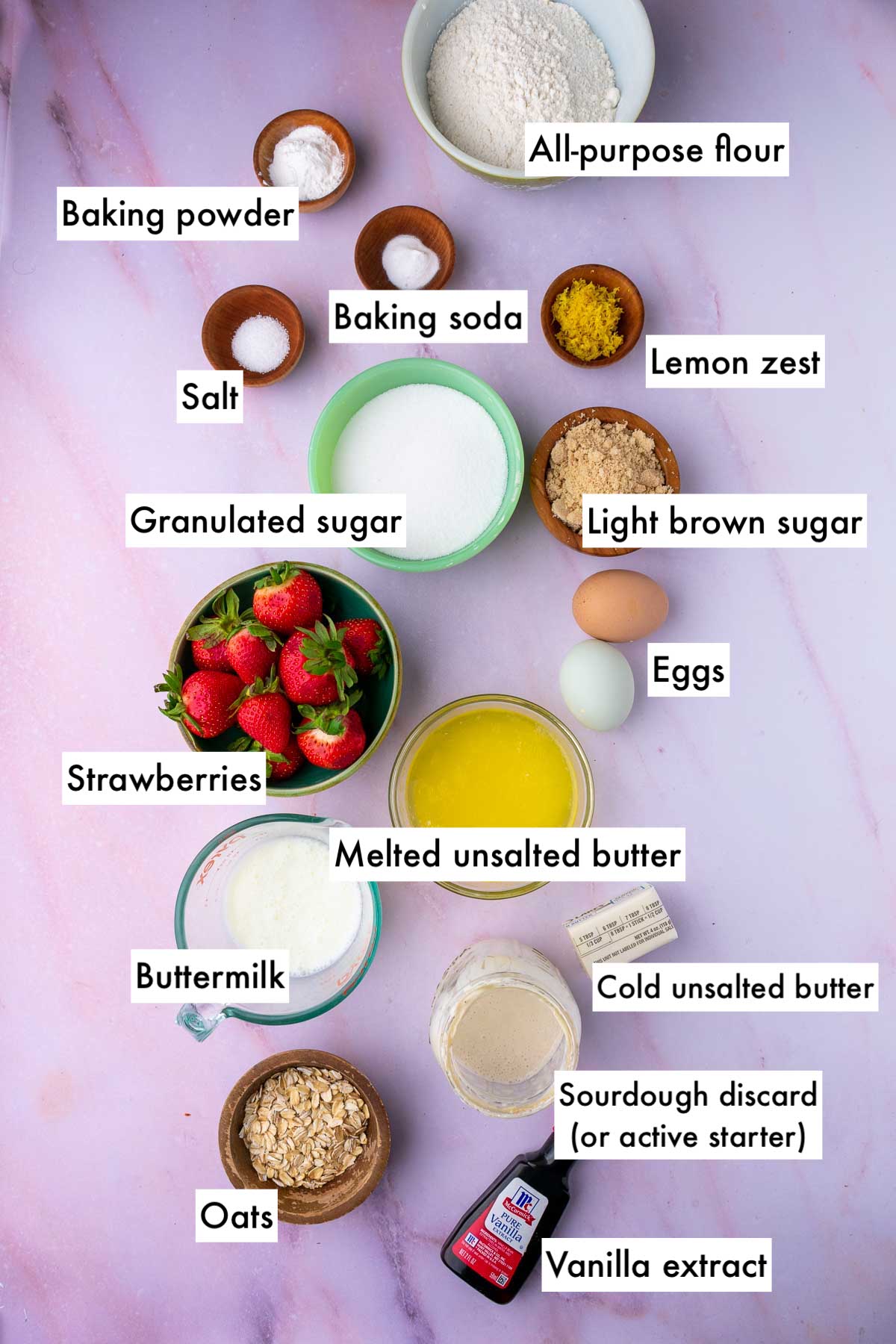 Labeled bowls of ingredients needed for Sourdough strawberry muffins including all-purpose flour, granulated sugar, light brown sugar, baking powder, baking soda, salt, eggs, strawberries, buttermilk, butter, vanilla, oats, and sourdough discard.