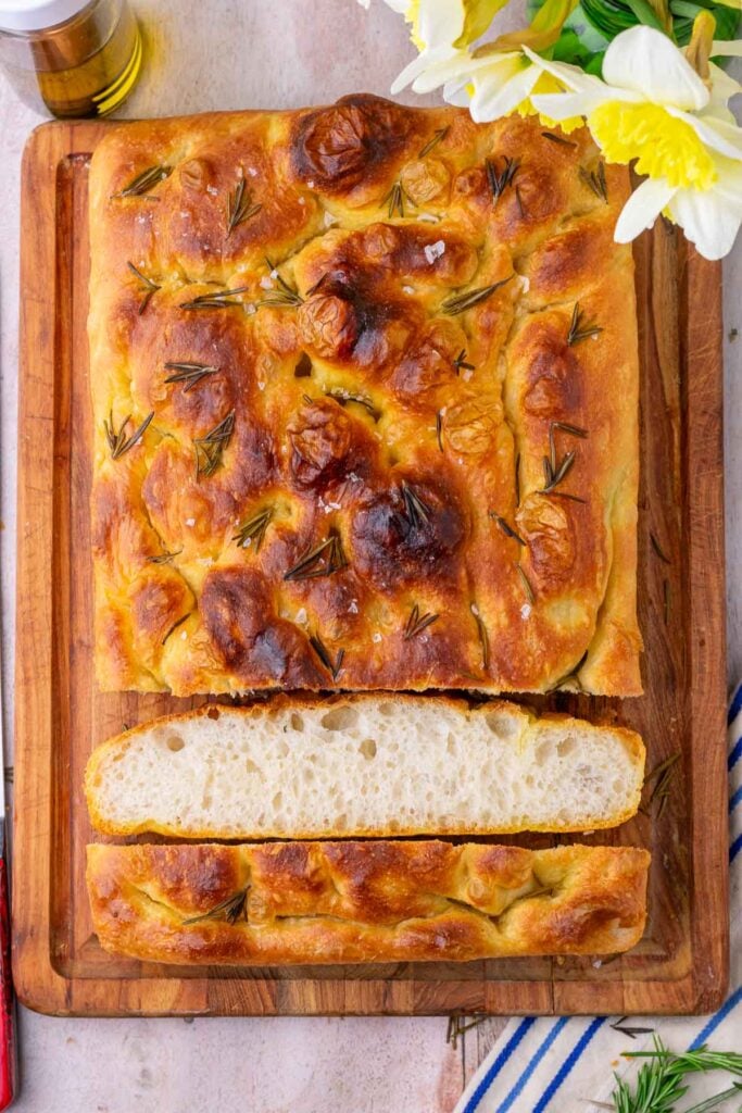 Sliced sourdough focaccia on a cutting board with rosemary, olive oil, and flowers.