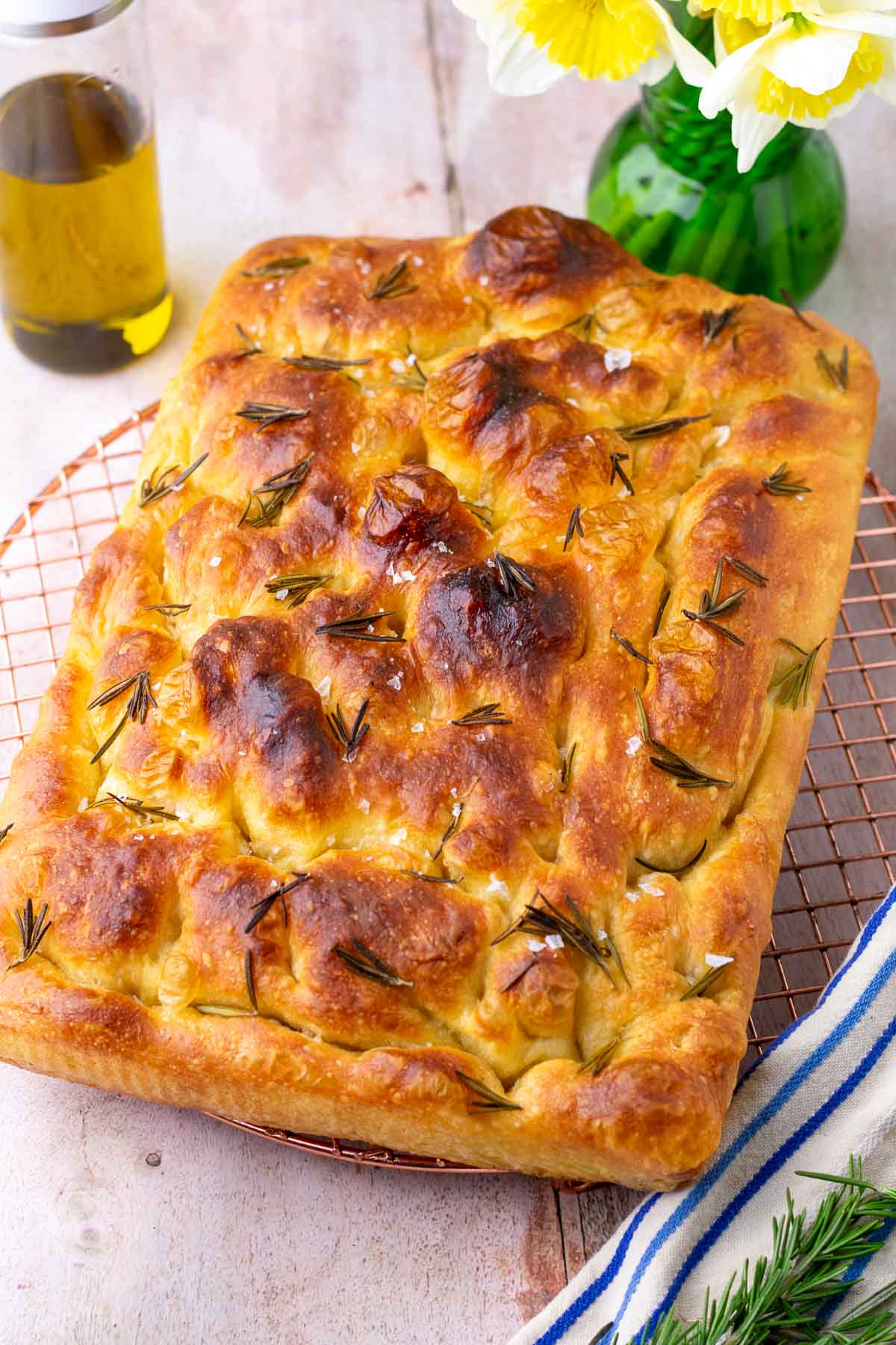 Baked sourdough focaccia on a wire rack with olive oil, rosemary, and flowers.