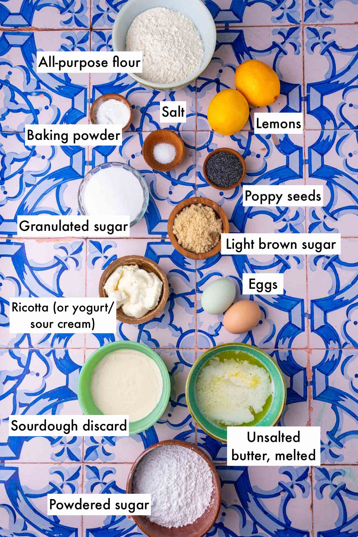 Labeled ingredients needed for sourdough lemon poppy seed muffins including all-purpose flour, lemons, poppy seeds, eggs, salt, baking powder, sourdough discard, melted butter, granulated sugar, light brown sugar, ricotta, and powdered sugar.