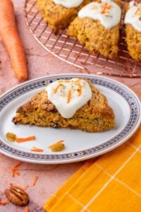 Sourdough carrot cake scone on a plate with more scones on a wire rack and carrots in the background.