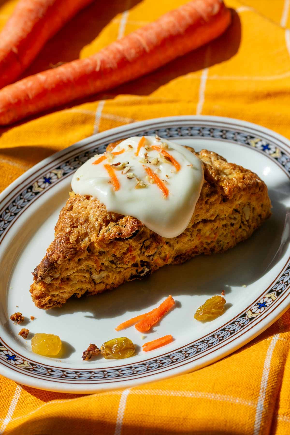 Sourdough carrot cake scone on a plate with cream cheese glaze and carrots in the background.
