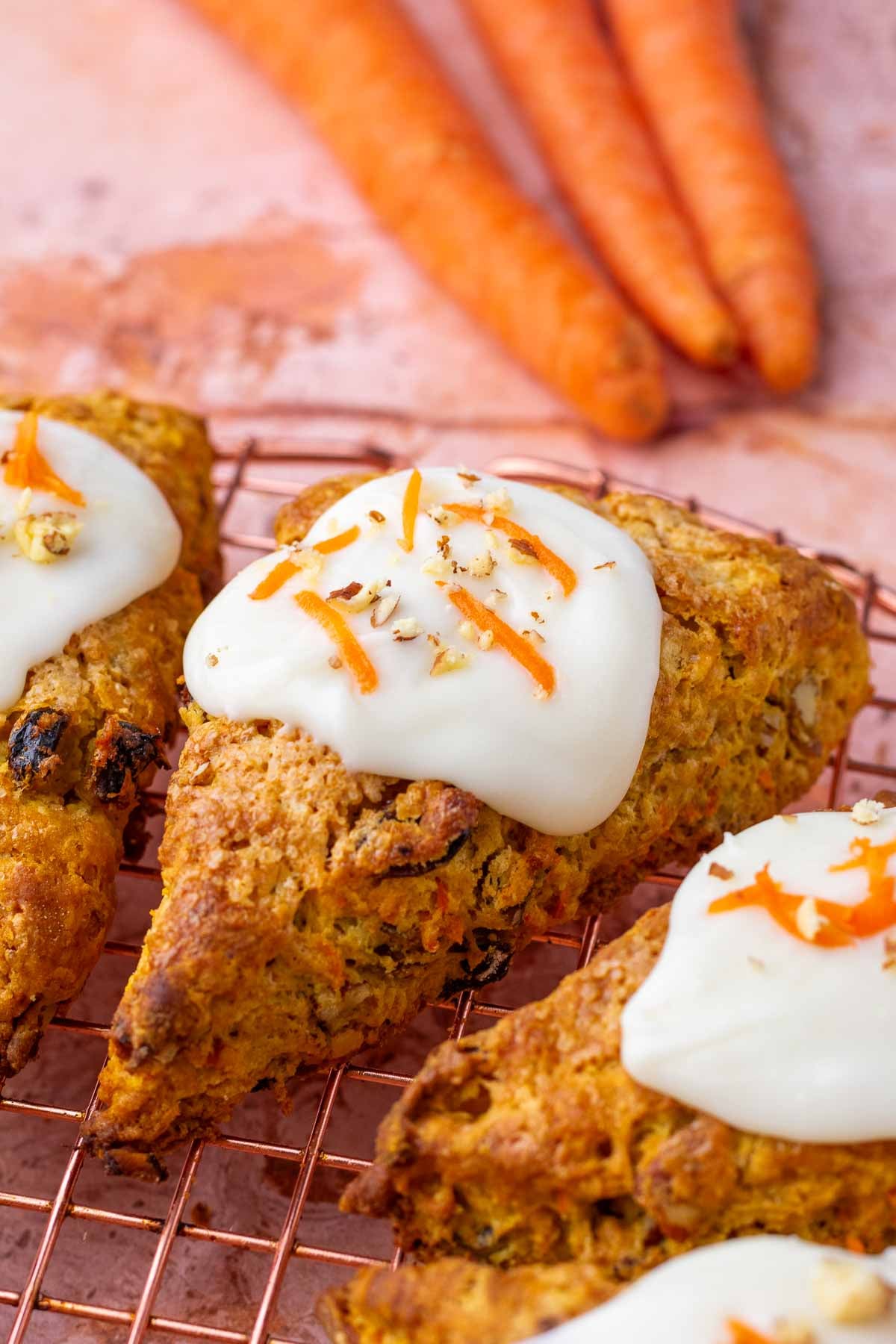  Close up of a sourdough carrot cake scone on a wire rack with cream cheese glaze and carrots in the background.