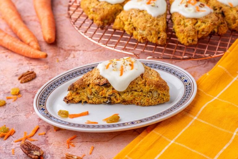 Sourdough carrot cake scone on a plate with cream cheese glaze on top and pecans, raisins, and carrots scattered around and more scones in the background on a wire rack.