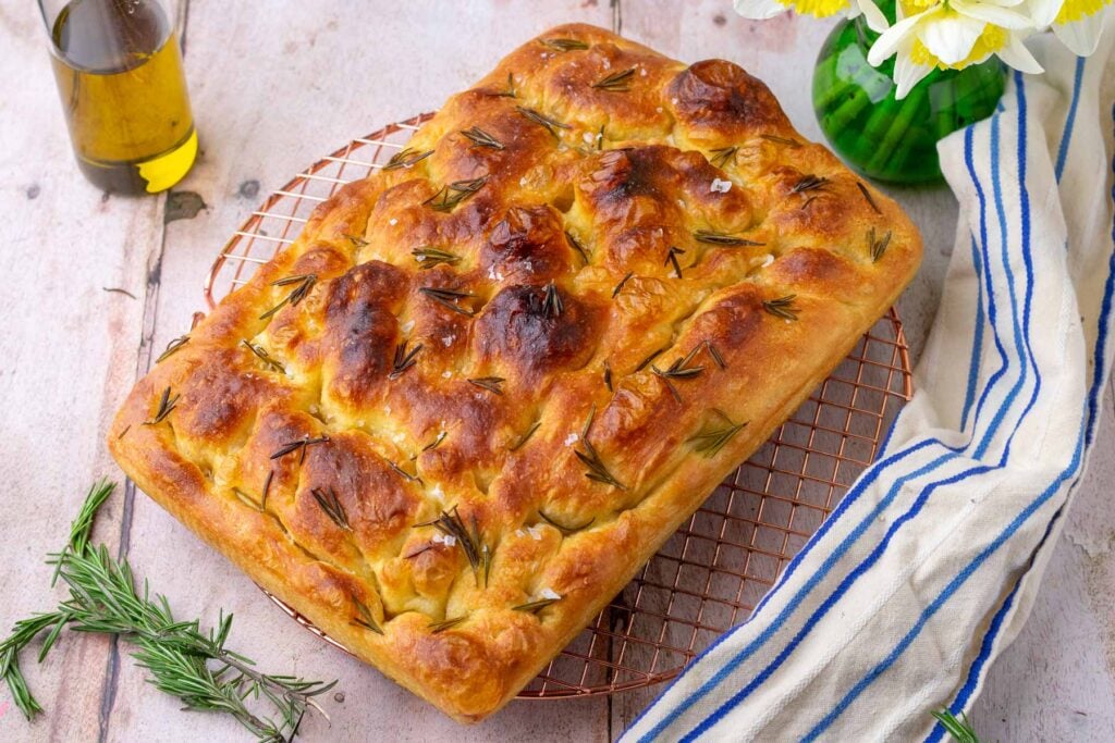 Sourdough focaccia on a wire rack with rosemary, olive oil, and flowers.