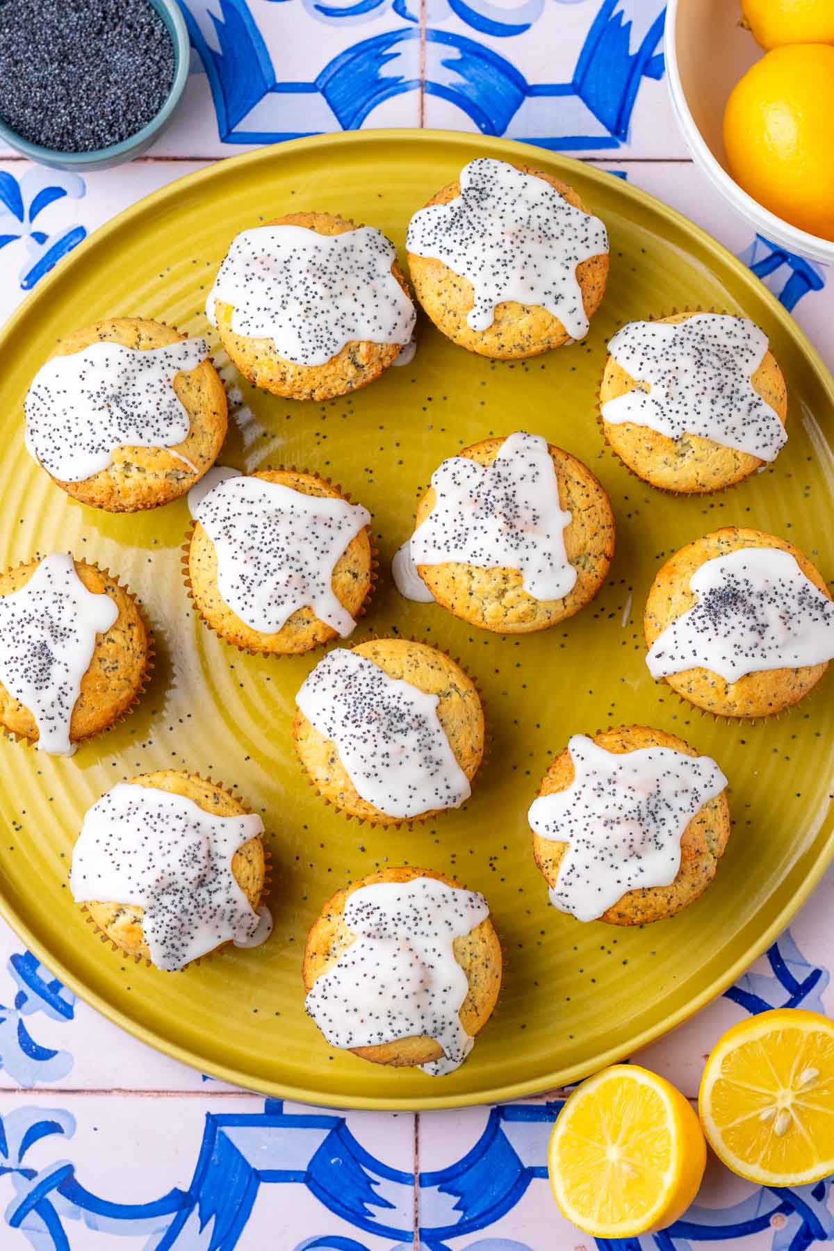Twelve sourdough lemon poppy seed muffins on a large platter with a sliced lemon, bowl of poppy seeds, and lemon bowl.