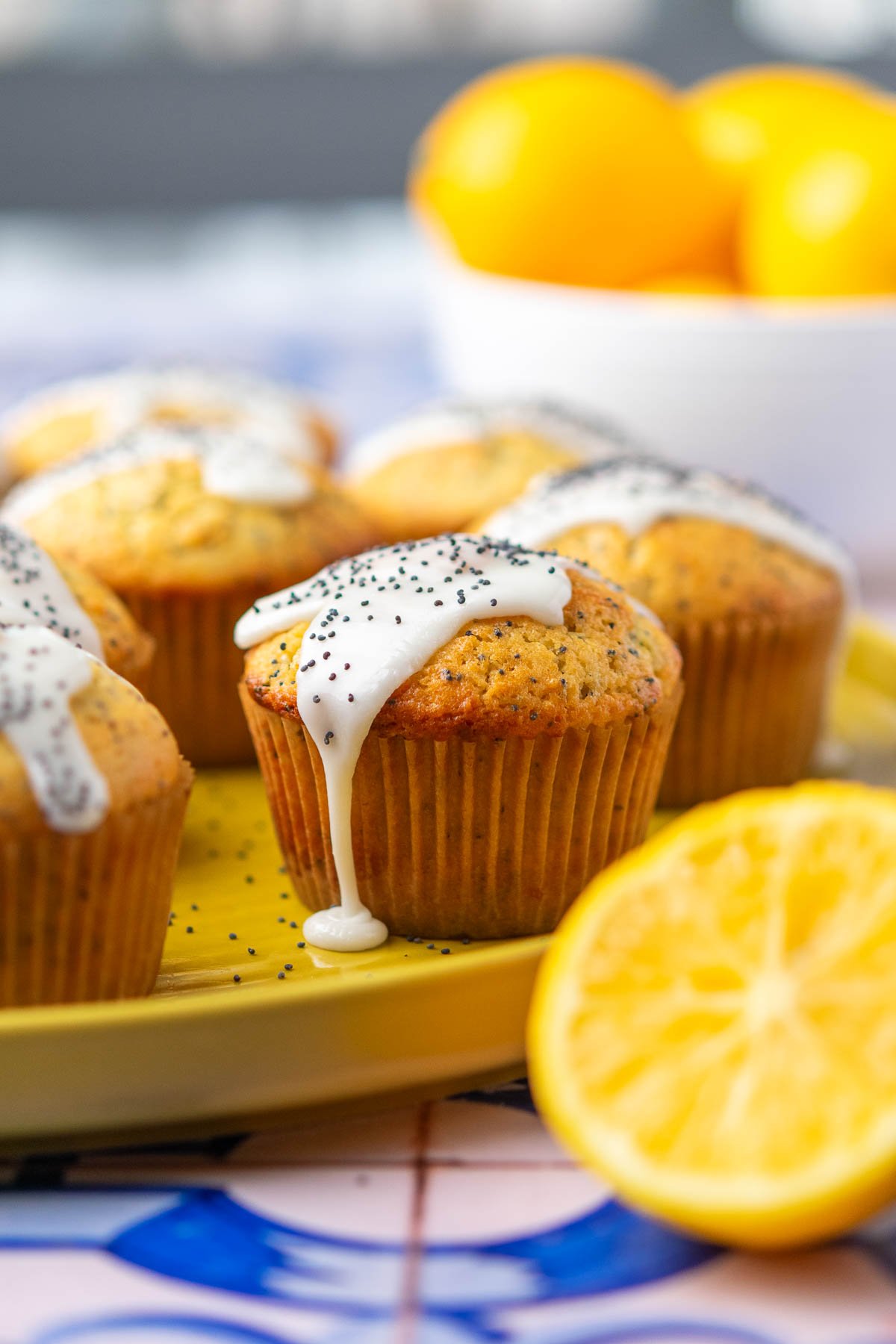 sourdough lemon poppy seed muffins on a plate with lemon glaze dripping, a sliced lemon in front, and a bowl of lemons in the back.