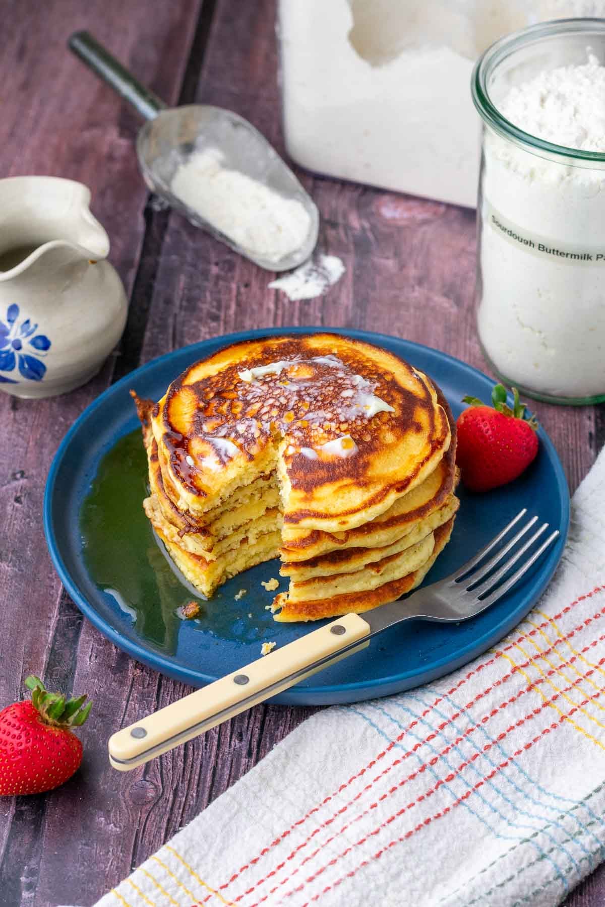 Bite taken out of a stack of sourdough buttermilk pancakes on a plate with maple syrup and sourdough buttermilk pancake mix in the background.