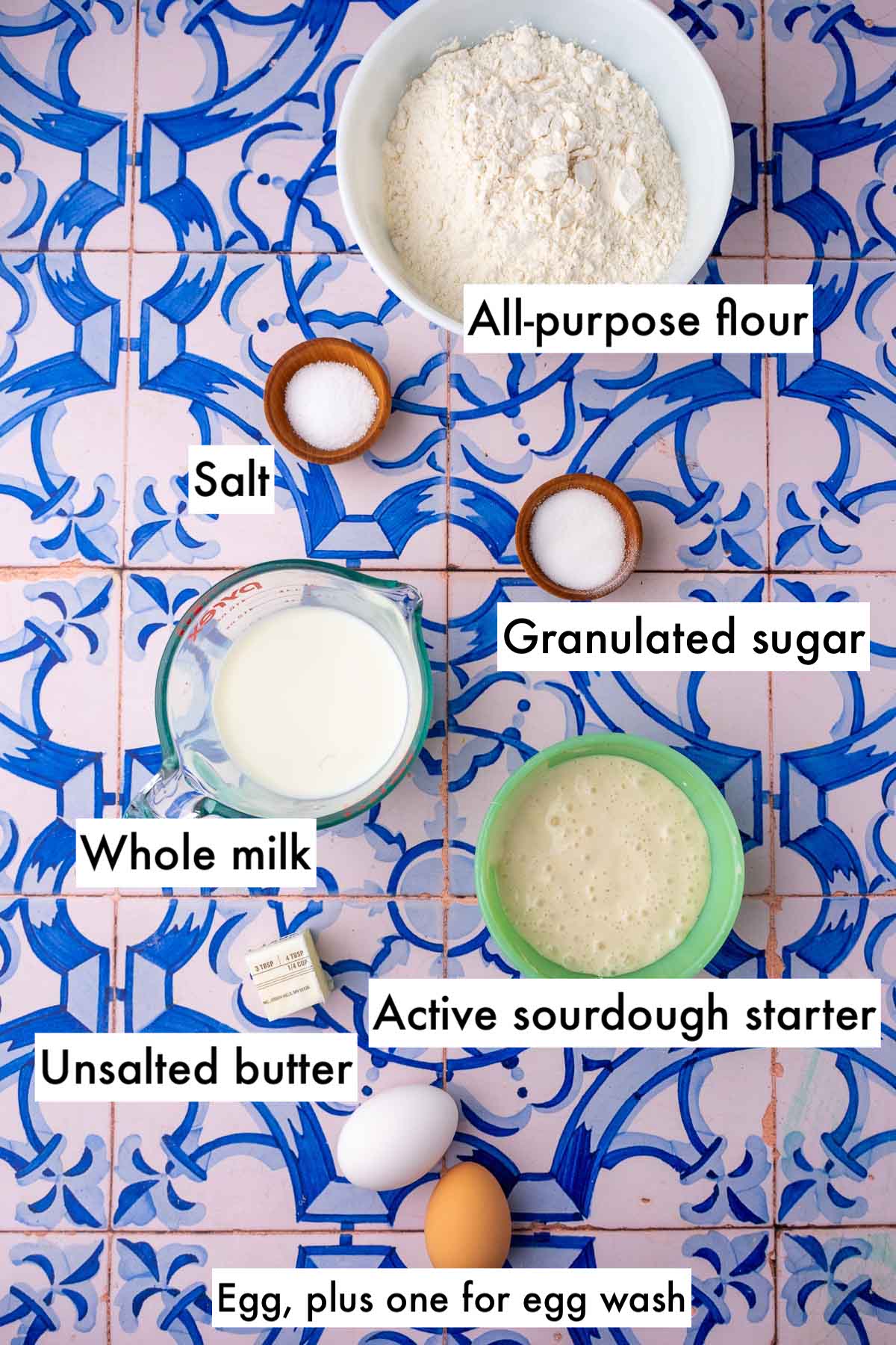 Labeled bowls of ingredients needed for sourdough hot dog buns including all-purpose flour, salt, sugar, whole milk, active sourdough starter, unsalted butter, and eggs.