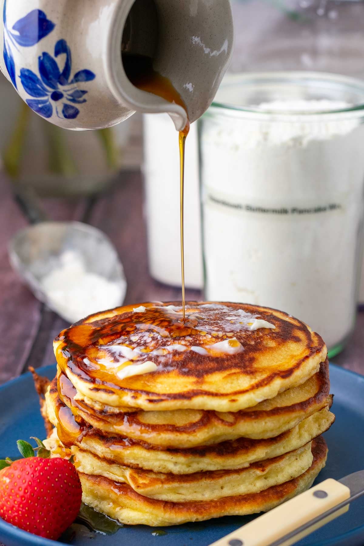 Pouring maple syrup onto a stack of sourdough buttermilk pancakes with a container of sourdough buttermilk pancake mix in the background.